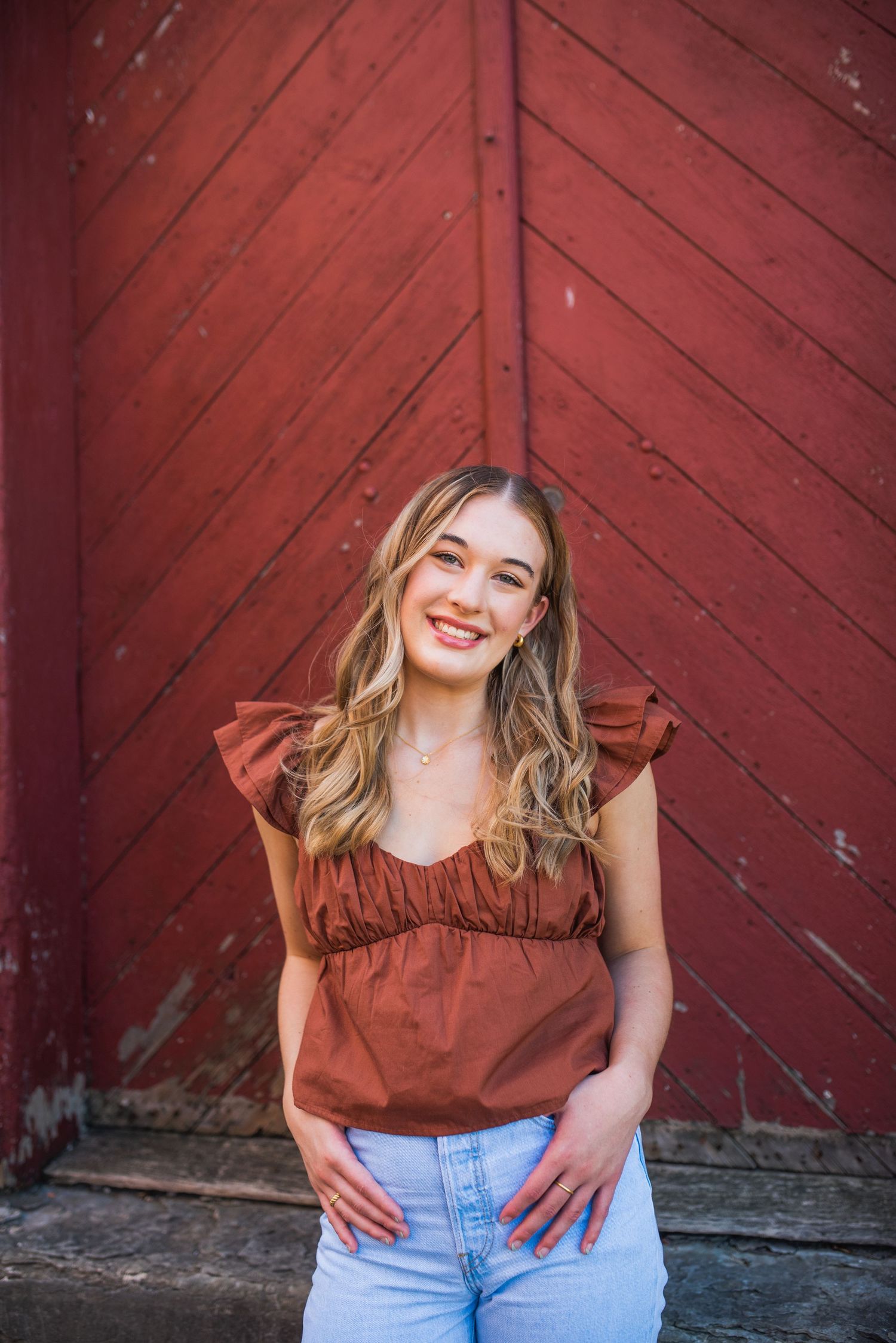 Portrait session against rustic red barn door wearing rust colored top and jeans.