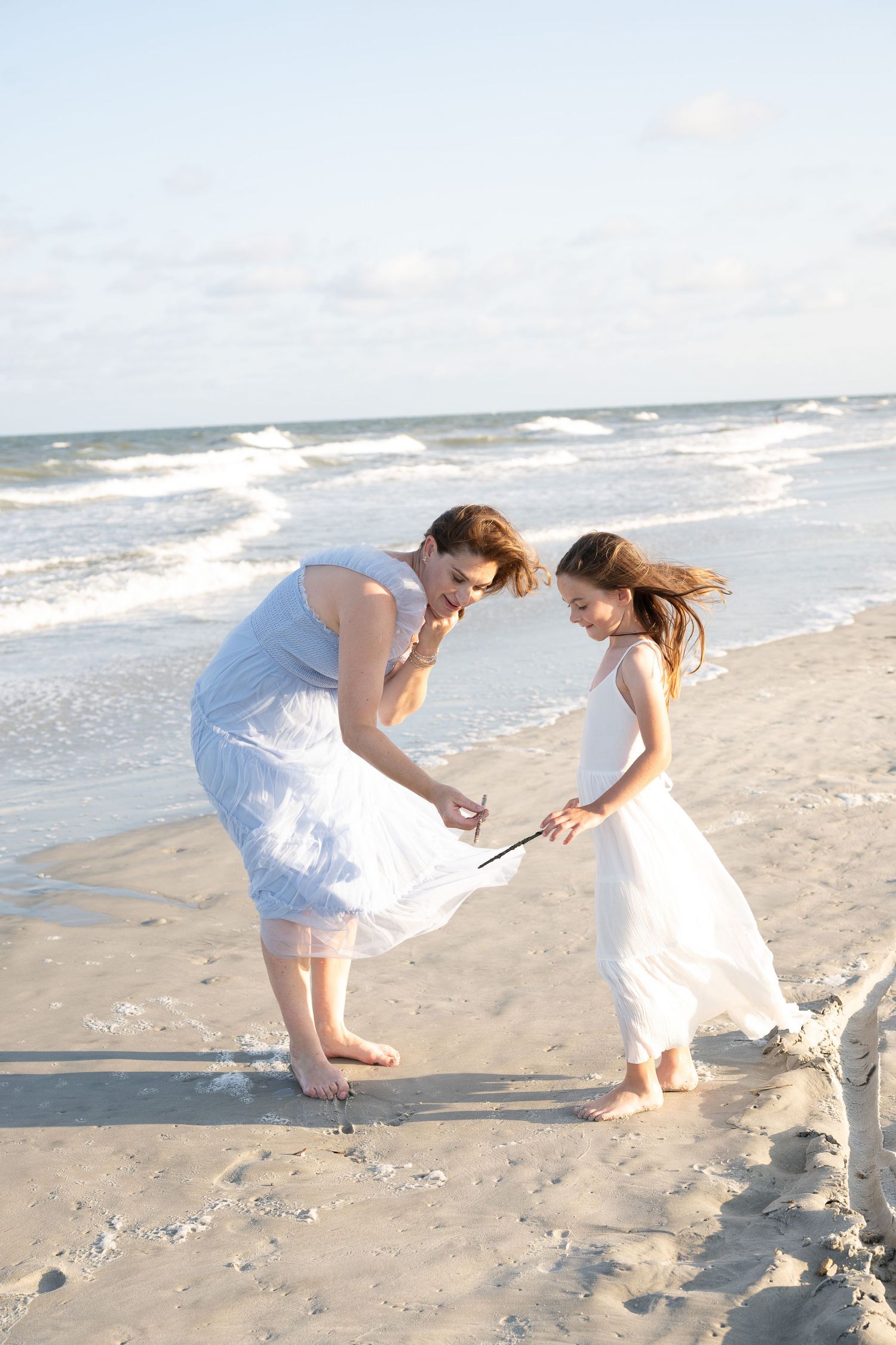 Two women in white dresses writing in the sand on a breezy beach with ocean waves in the background.