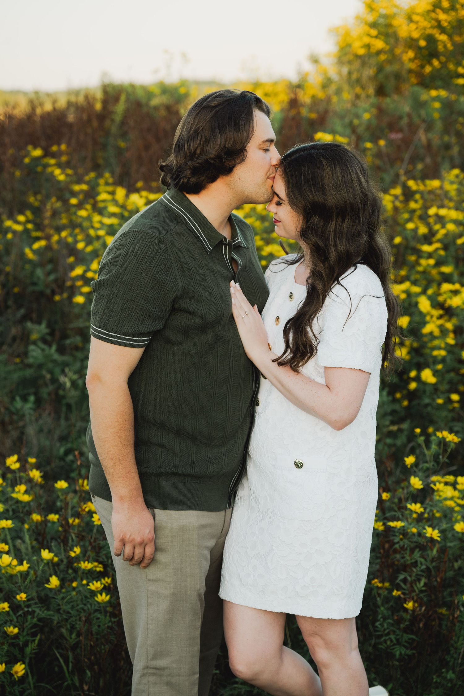 Couple sharing a romantic kiss in a field of yellow wildflowers, man in green polo and woman in white dress.