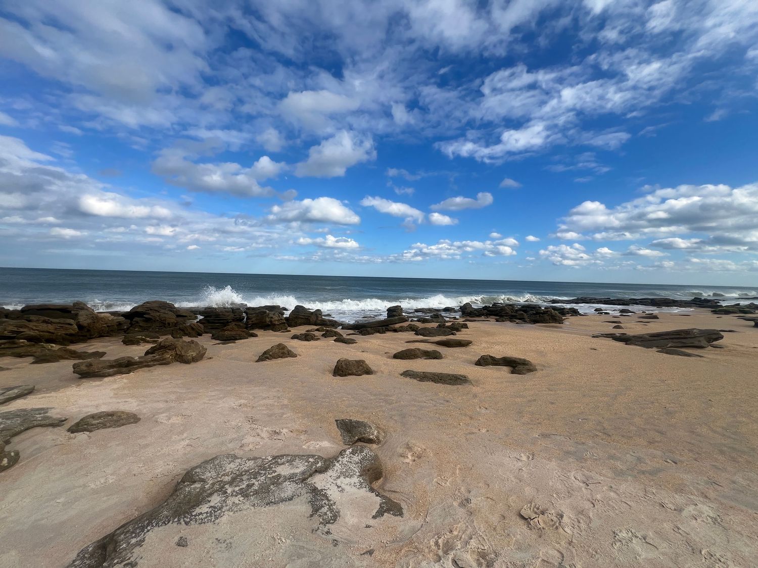 Rocky beach with crashing waves under a blue sky with white clouds, sandy shore with scattered boulders near the ocean.