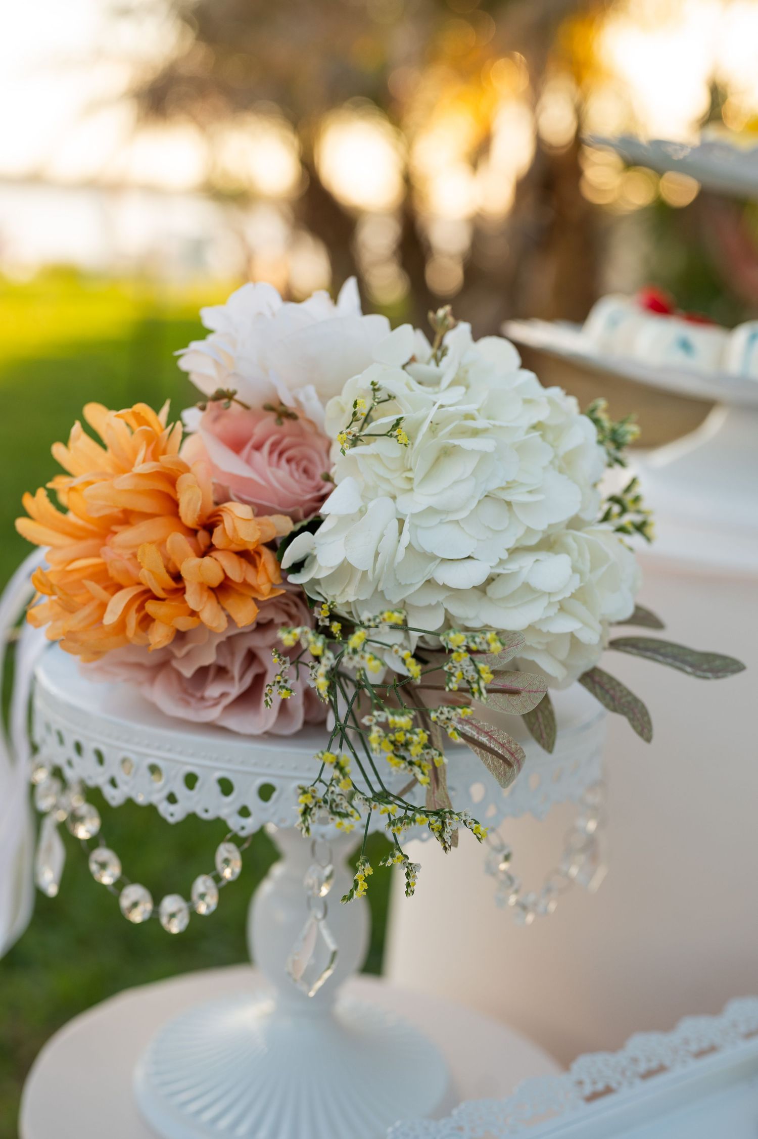 White cake stand adorned with orange dahlia, blush roses, and white hydrangea floral arrangement at outdoor wedding.
