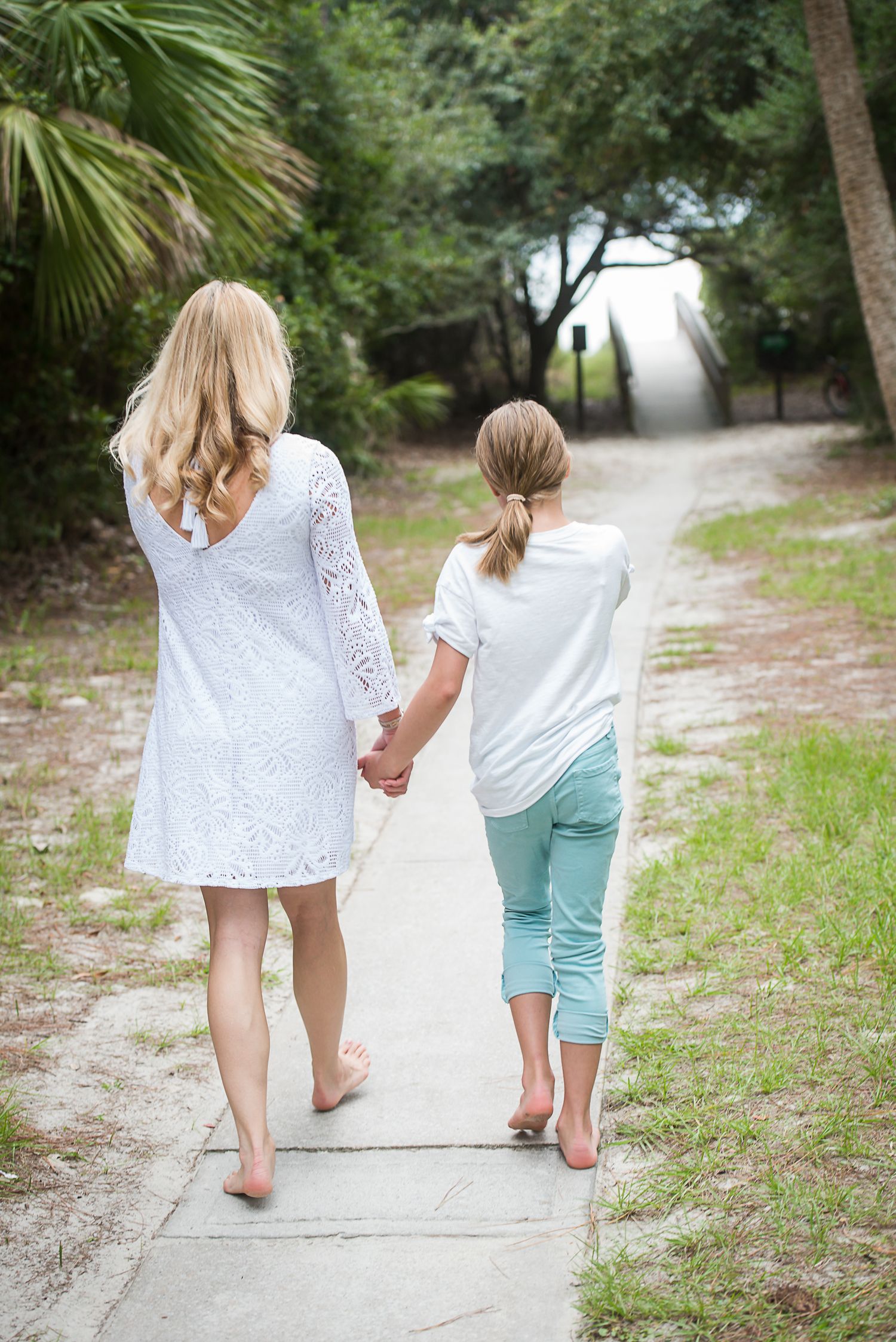 Mother and child walking barefoot together on sandy beach path holding hands. Hammock Beach engagement session.