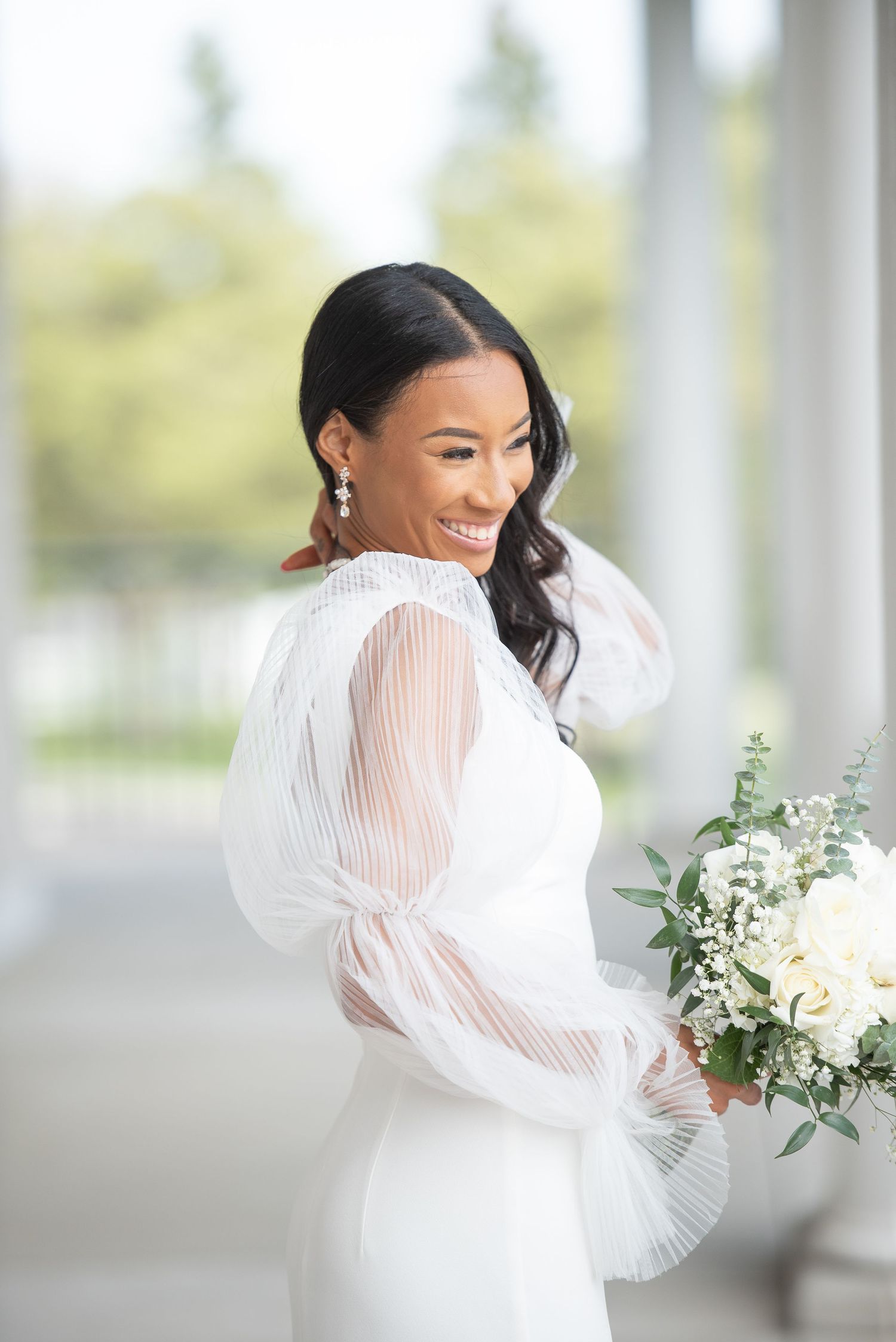 Bride in elegant white dress with sheer sleeves holding bouquet of white flowers.