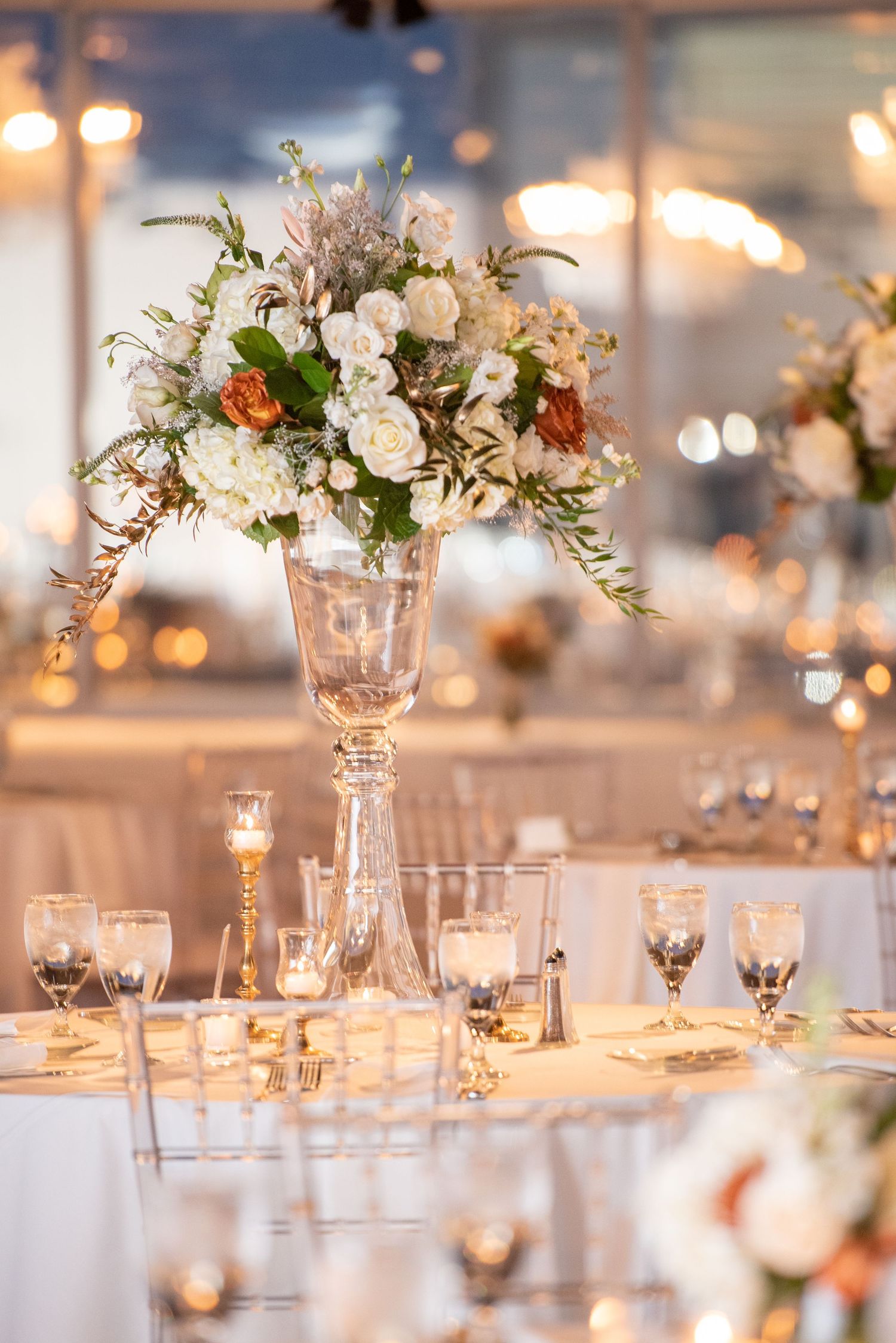 Elegant wedding centerpiece with white roses and greenery in tall crystal vase on table with glowing candlelight.