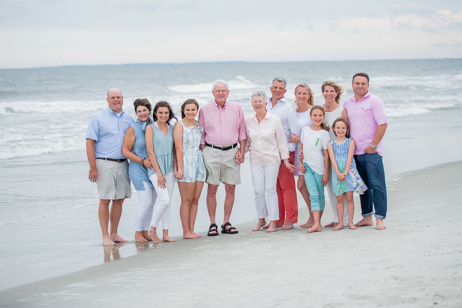 Hilton Head extended Family group wearing white and pastel colors stands together on a sandy beach at sunset.