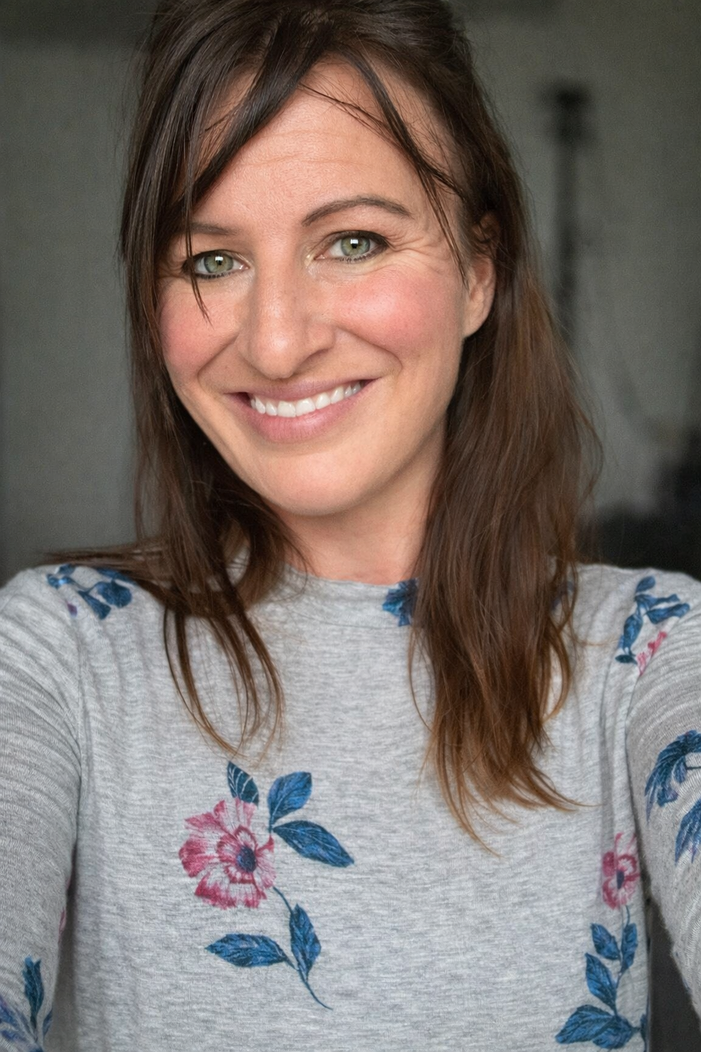 Smiling woman with brown hair and green eyes wearing a gray floral embroidered top in a bright indoor setting.