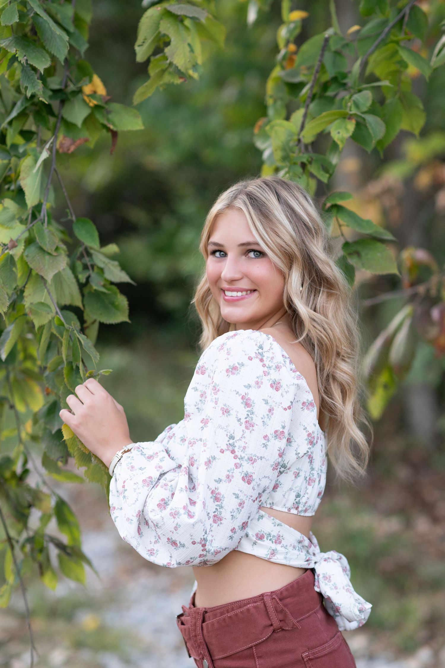 Young blonde woman in floral crop top and burgundy jeans smiling outdoors among green leaves.