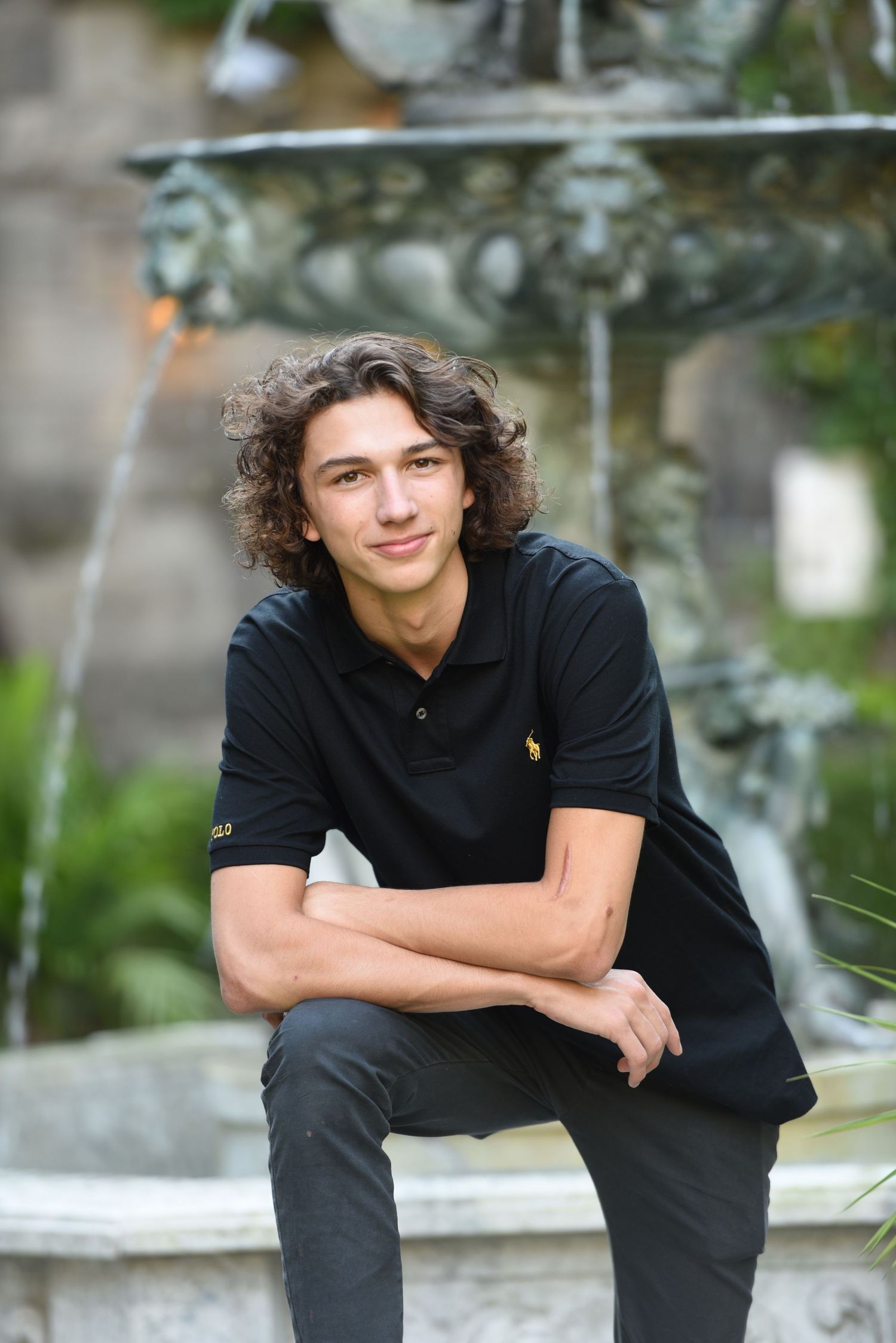Young person in black polo shirt sits casually on stone ledge with curly hair and relaxed pose in outdoor setting.