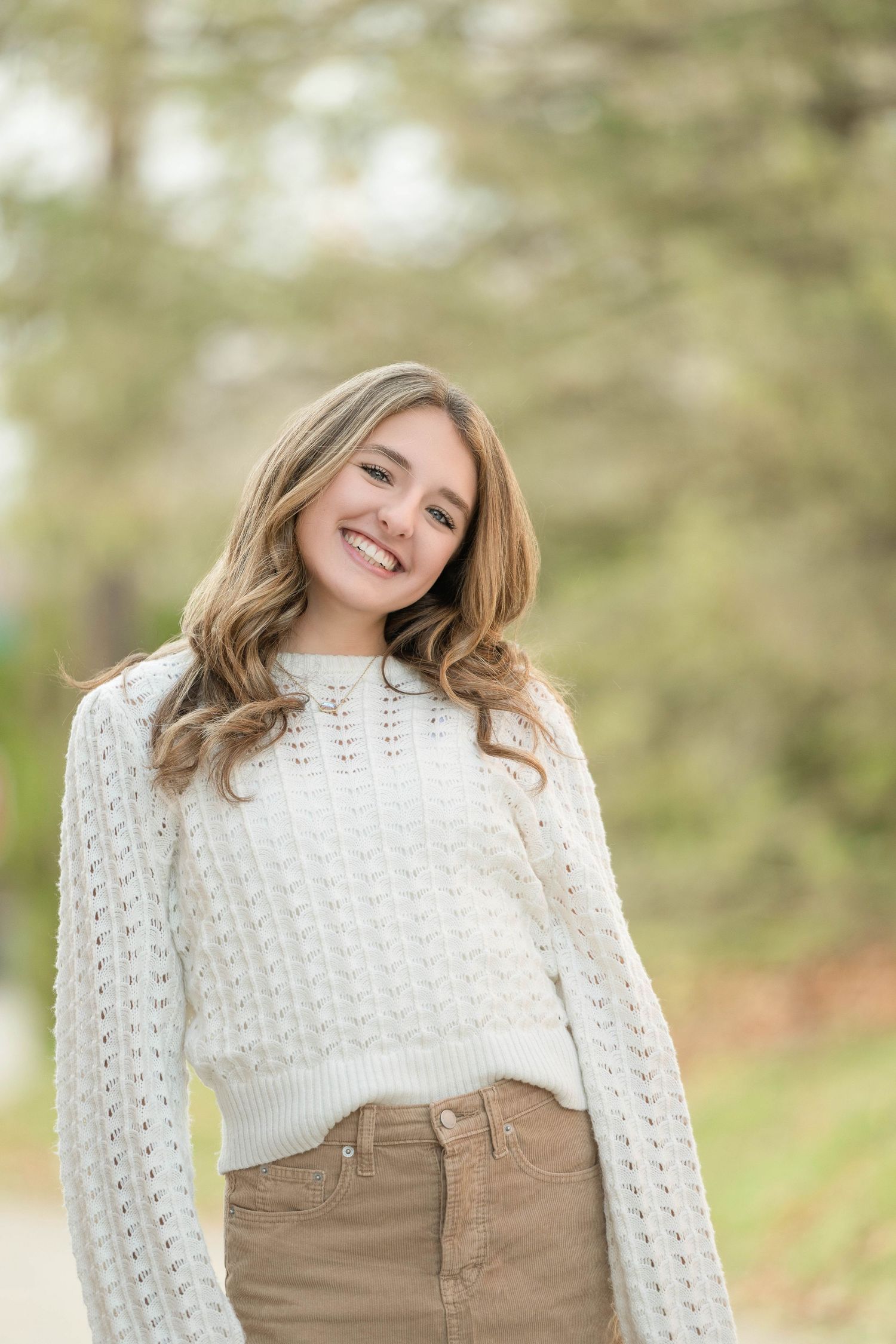 Young woman wearing a cozy white cable knit sweater and khaki pants smiles outdoors in natural lighting.