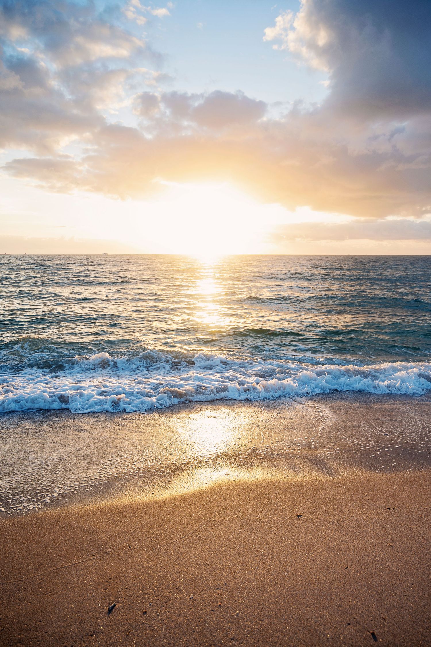 Golden sunset illuminates waves crashing on sandy beach with dramatic clouds and ocean horizon in warm evening light.