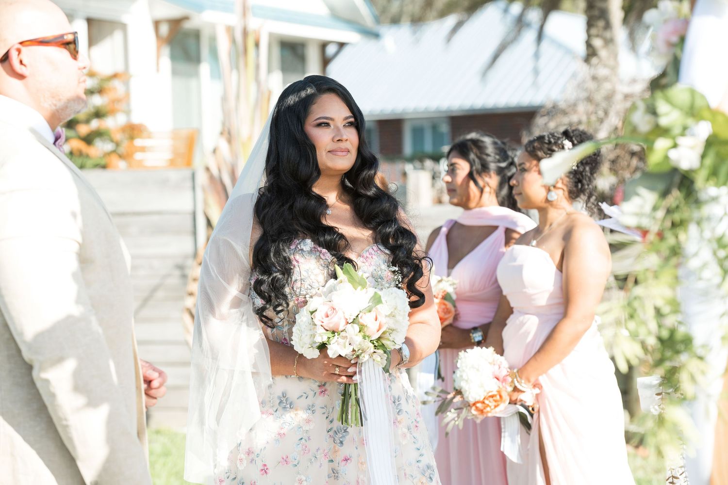 Bride in floral gown holding white bouquet stands with bridesmaids in pink dresses at outdoor wedding ceremony.