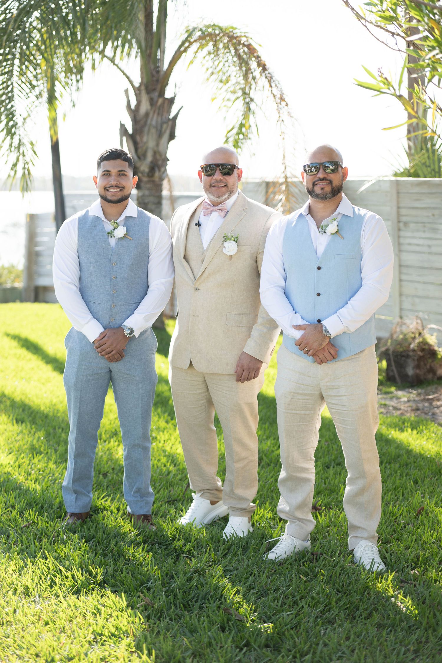 Three men in wedding attire pose outdoors on green grass, wearing vests and boutonnieres near a palm tree.