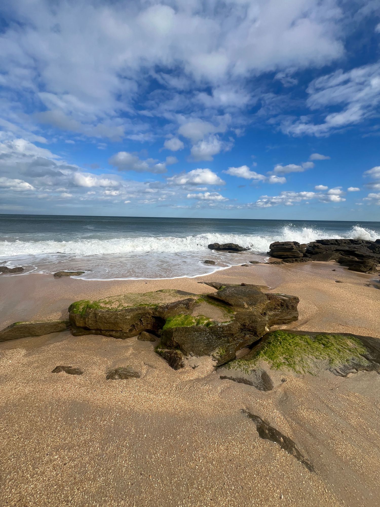 Rocky beach shoreline with moss-covered stones, crashing waves, golden sand, and a blue sky with fluffy white clouds.
