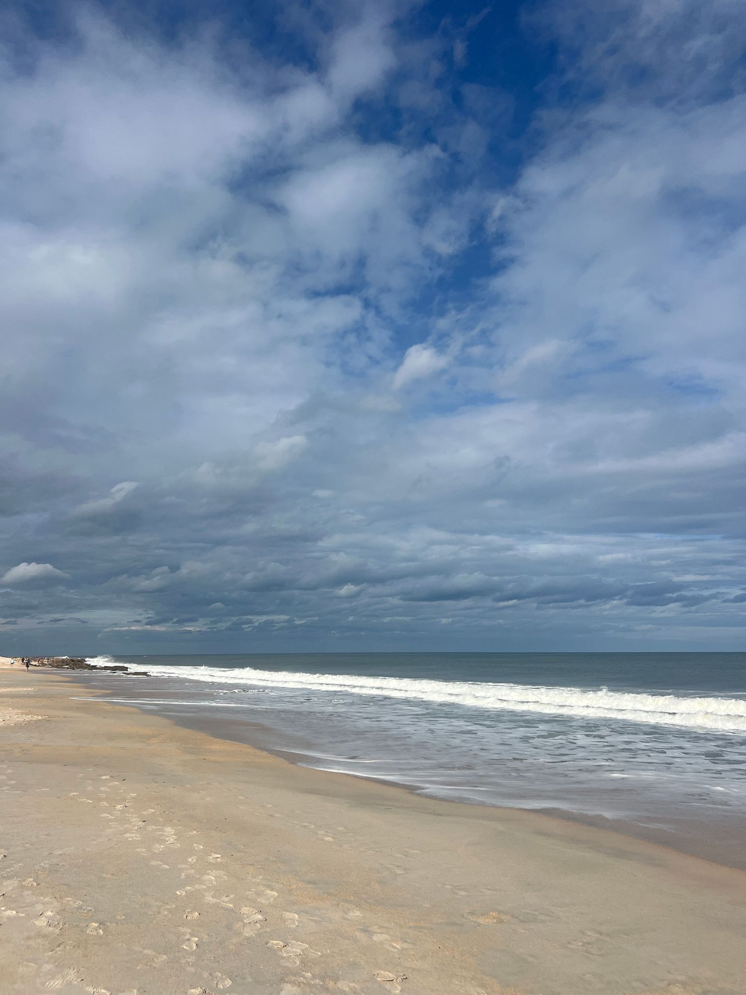 Sandy beach with gentle waves under a dramatic cloudy blue sky stretching to the horizon on a calm day.