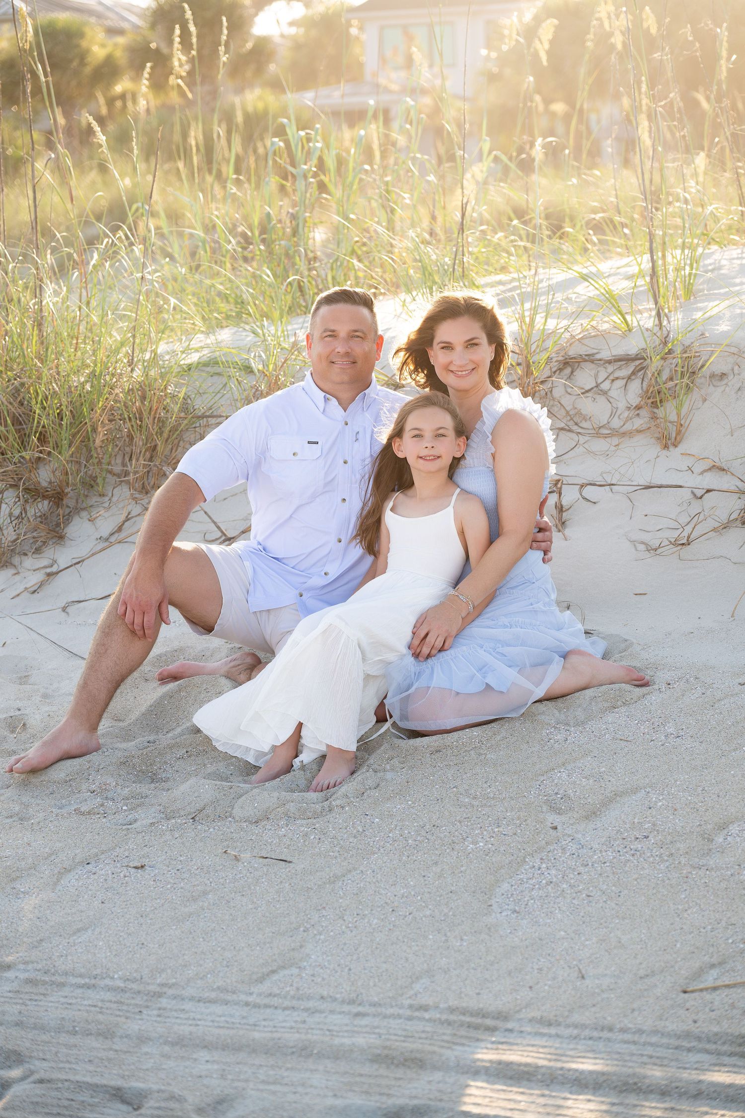 Family of three sitting on sandy beach dunes, wearing white and blue outfits, smiling warmly in golden sunlight.