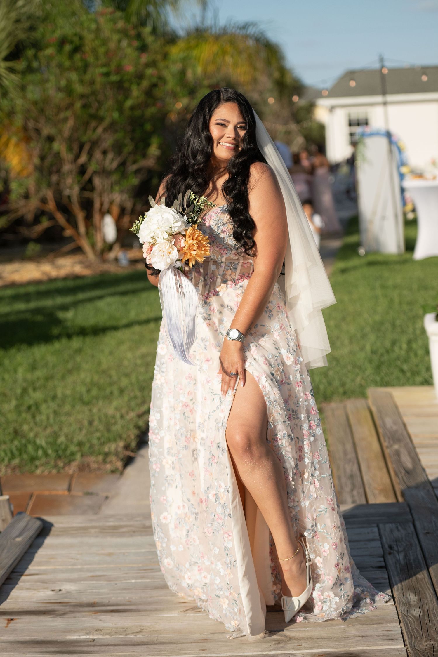 Smiling bride in floral lace gown with veil holds bouquet on wooden walkway outdoors.