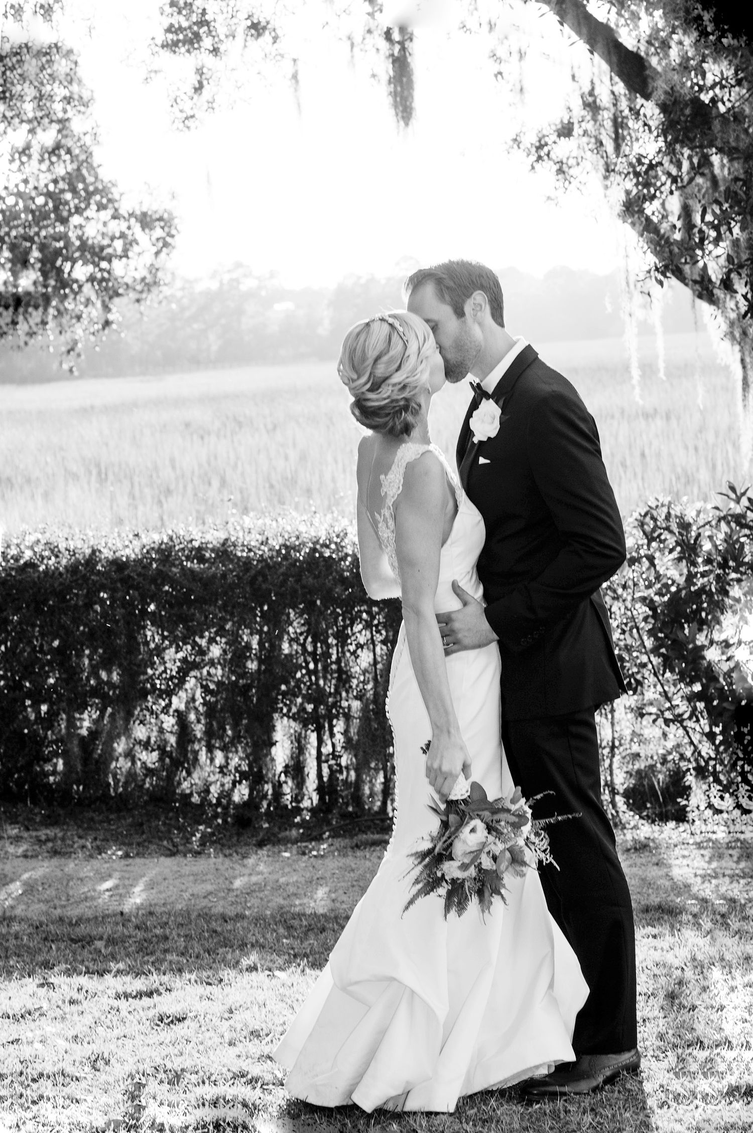 A black and white wedding photo captures a romantic moment between newlyweds under a canopy of trees.