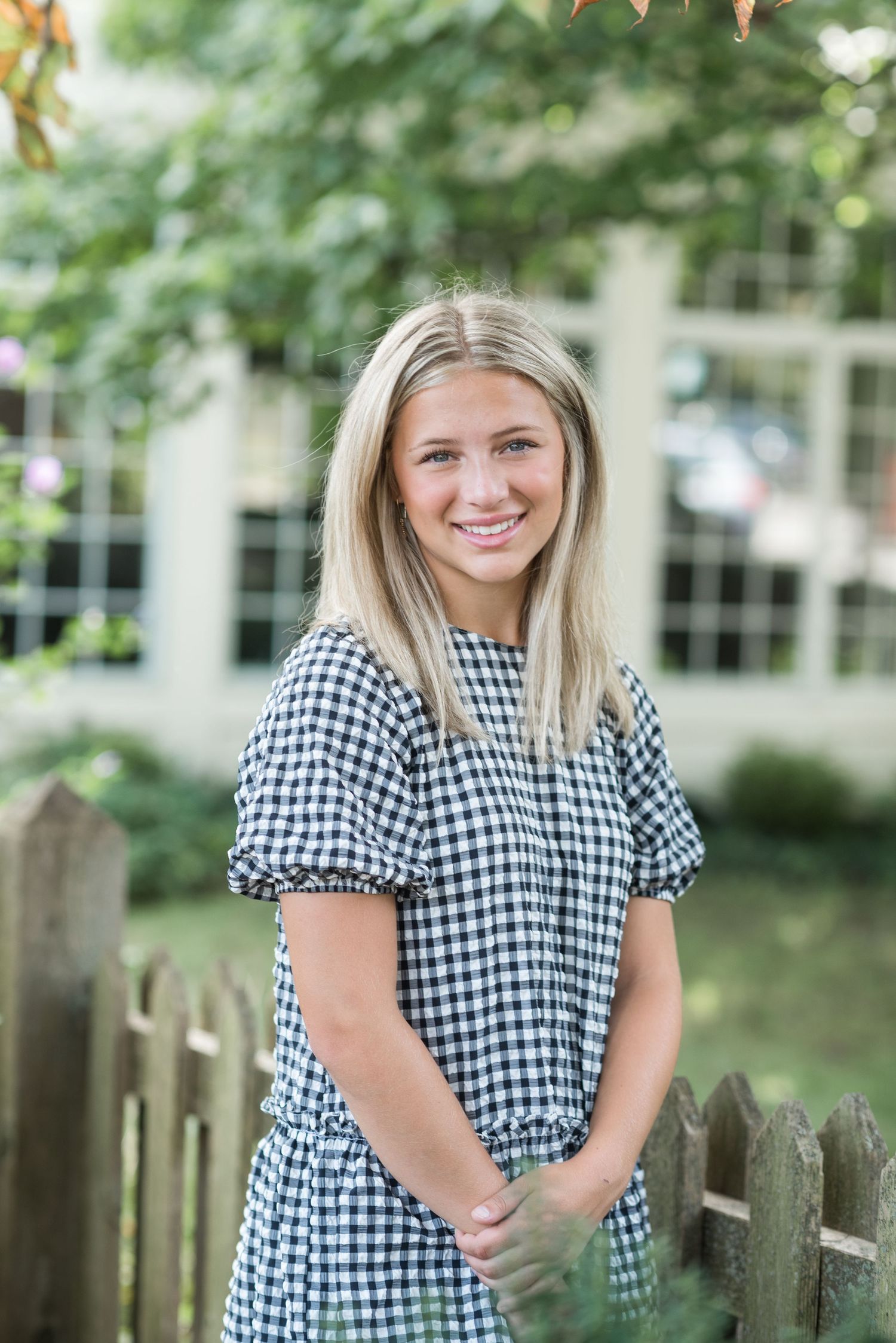 A Palm Coast senior portrait wearing a black and white gingham print top poses outdoors against a wooden fence with trees in background.