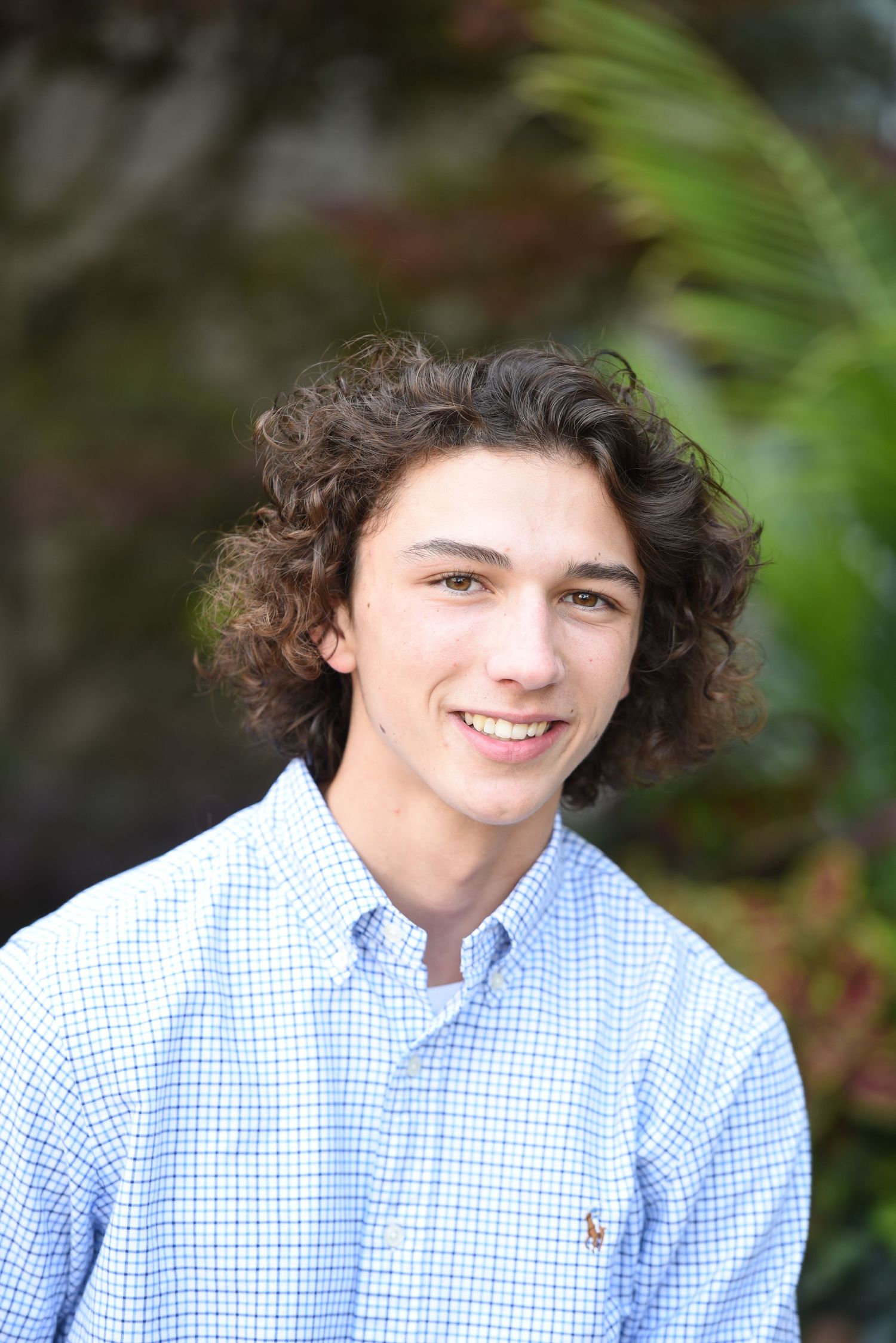 Portrait of someone with curly dark hair wearing a light blue checkered button-down shirt against a blurred green background.