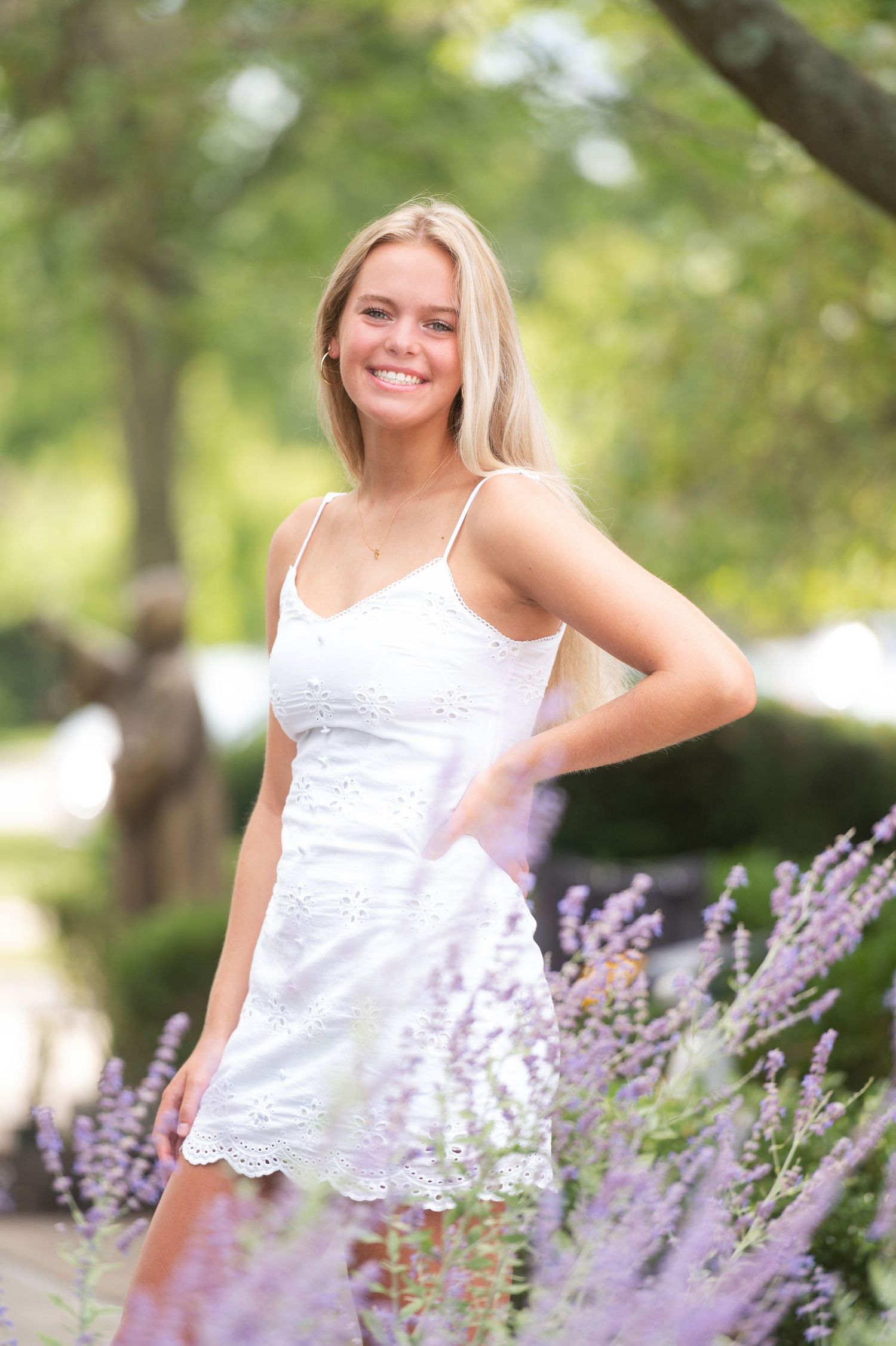 A woman in a white lace sundress poses in a garden with blooming lavender flowers in the foreground.