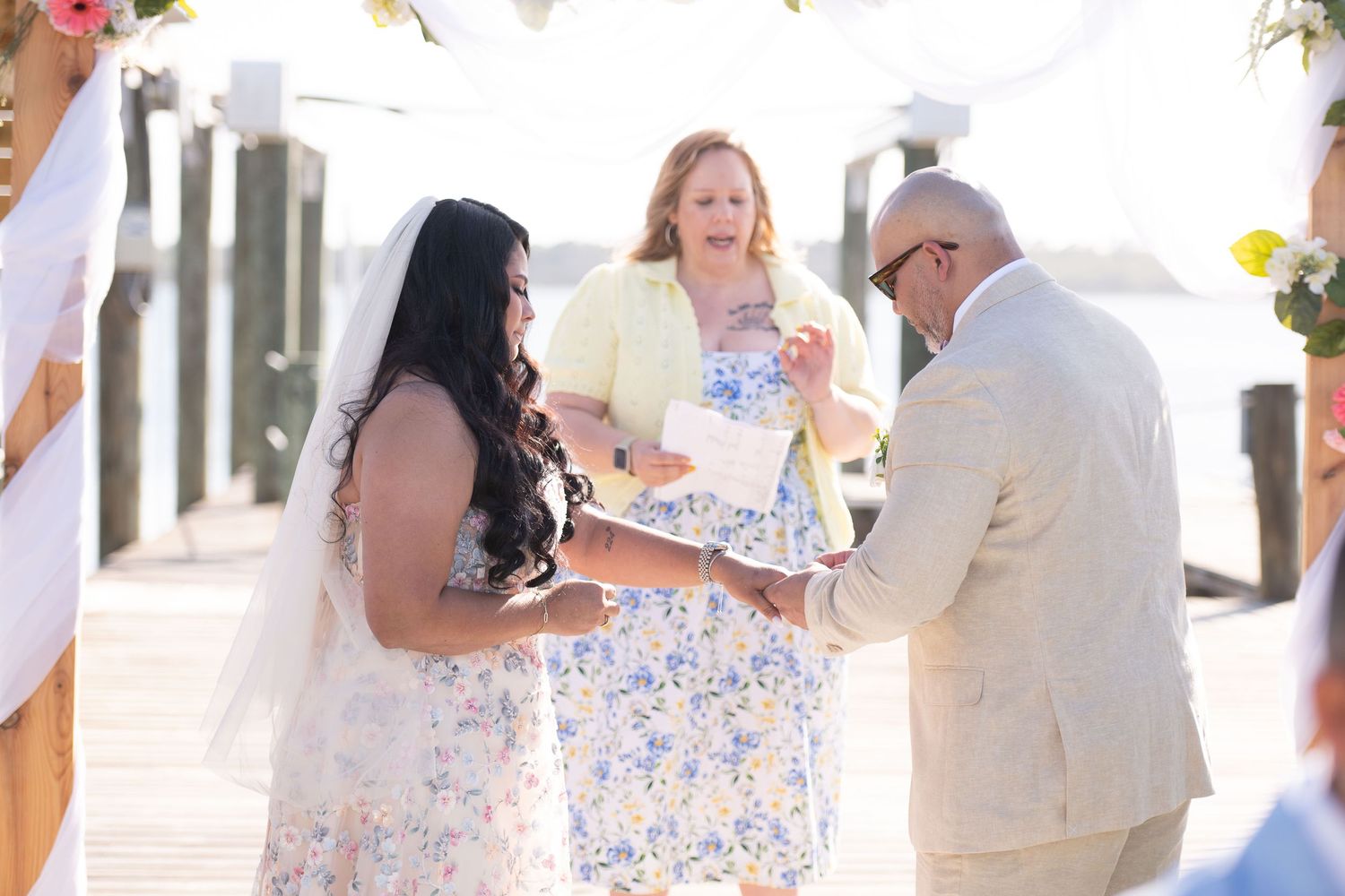 Outdoor waterfront wedding ceremony with bride in floral dress, groom in tan suit, and officiant exchanging vows on a sunny dock.