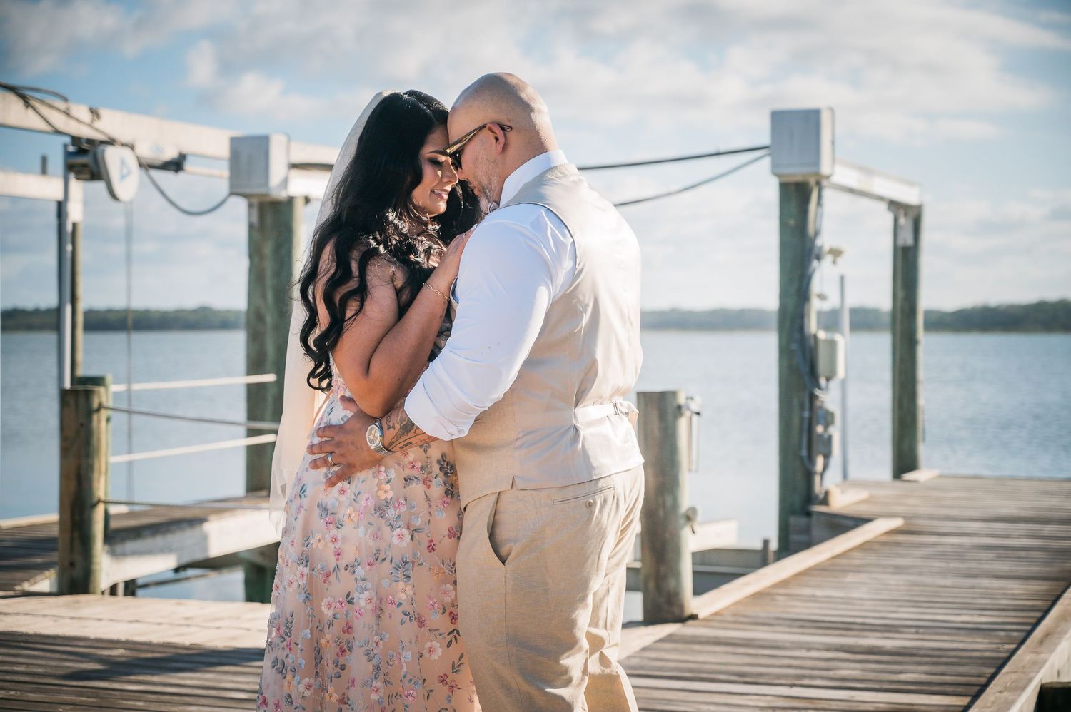 Couple sharing a romantic embrace on a wooden dock by the water, woman in floral dress and man in tan vest.