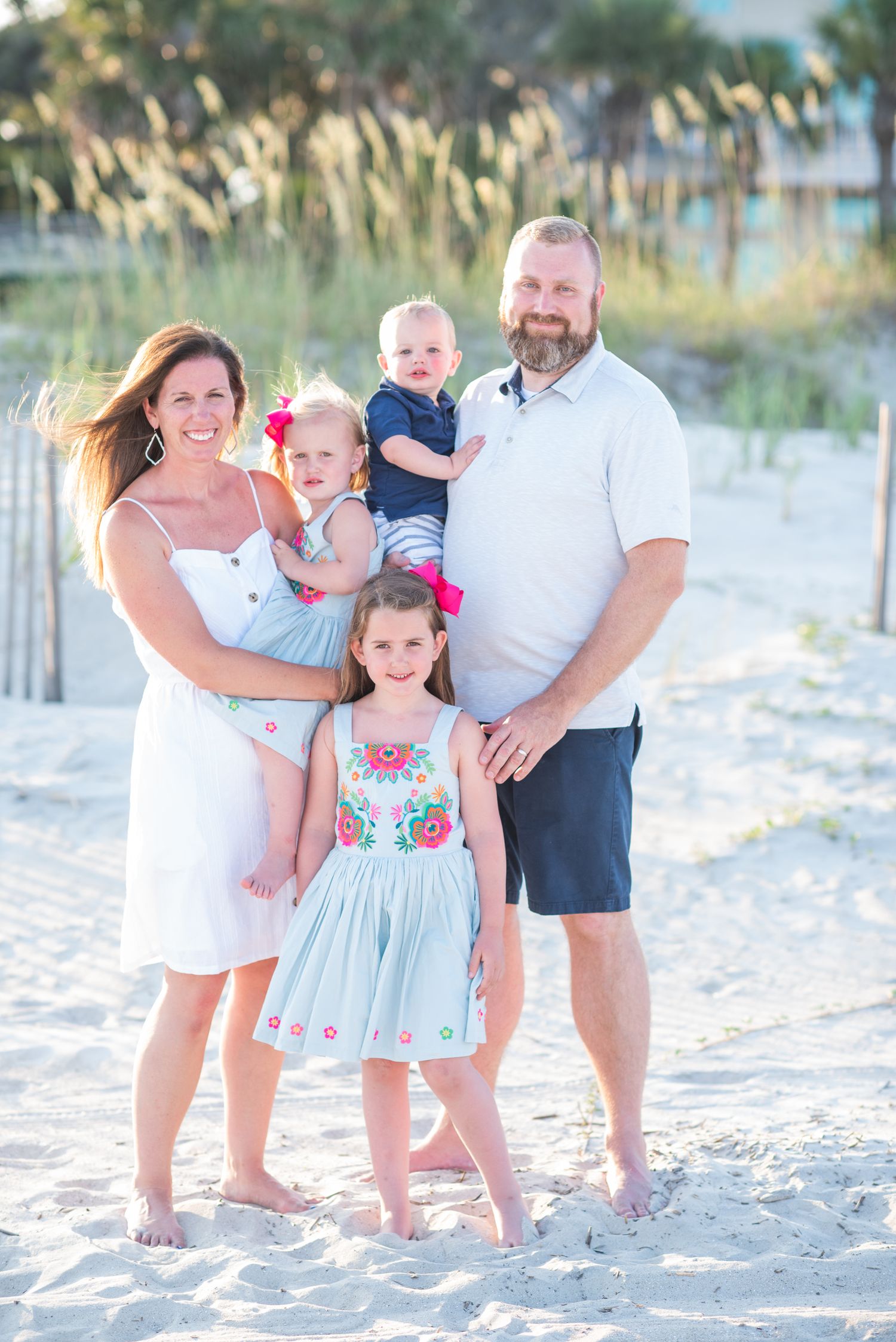 Family of five poses on white sand beach, parents holding toddlers with older daughter in floral dress smiling in front.