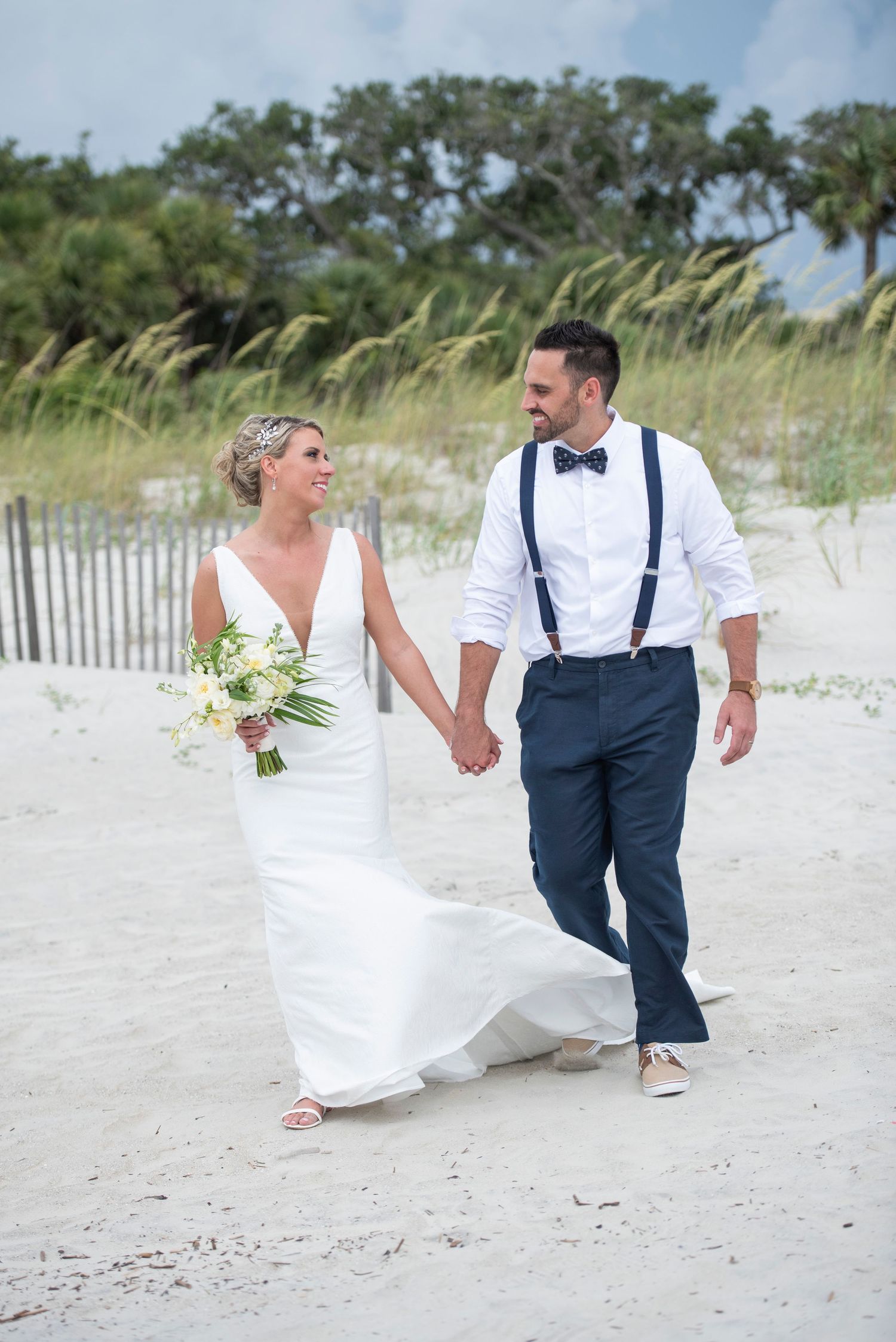 Bride and groom walk hand-in-hand on a sandy beach, she in a white gown, he in navy pants and suspenders.