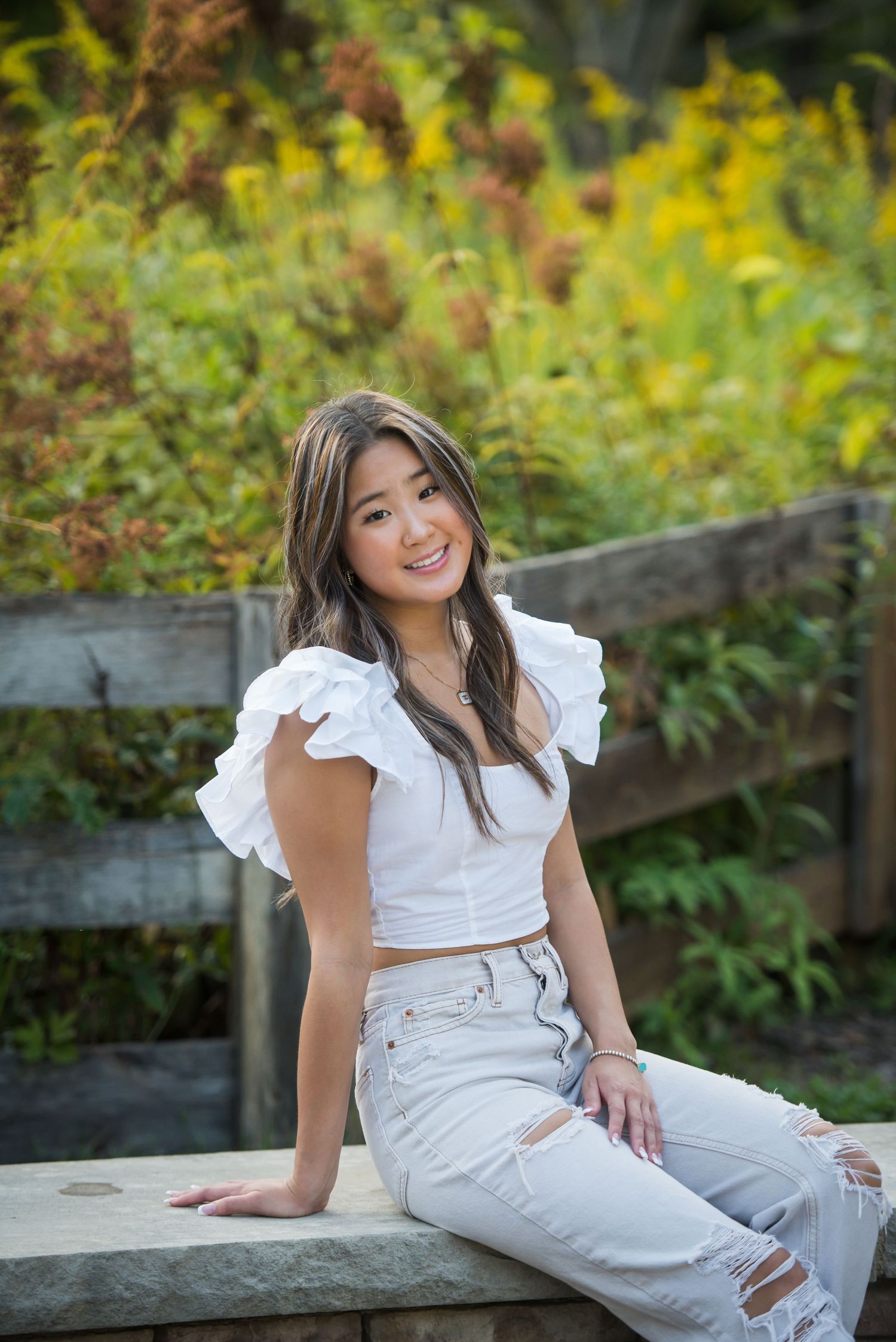 Person in white ruffled outfit poses on wooden bench surrounded by yellow wildflowers.