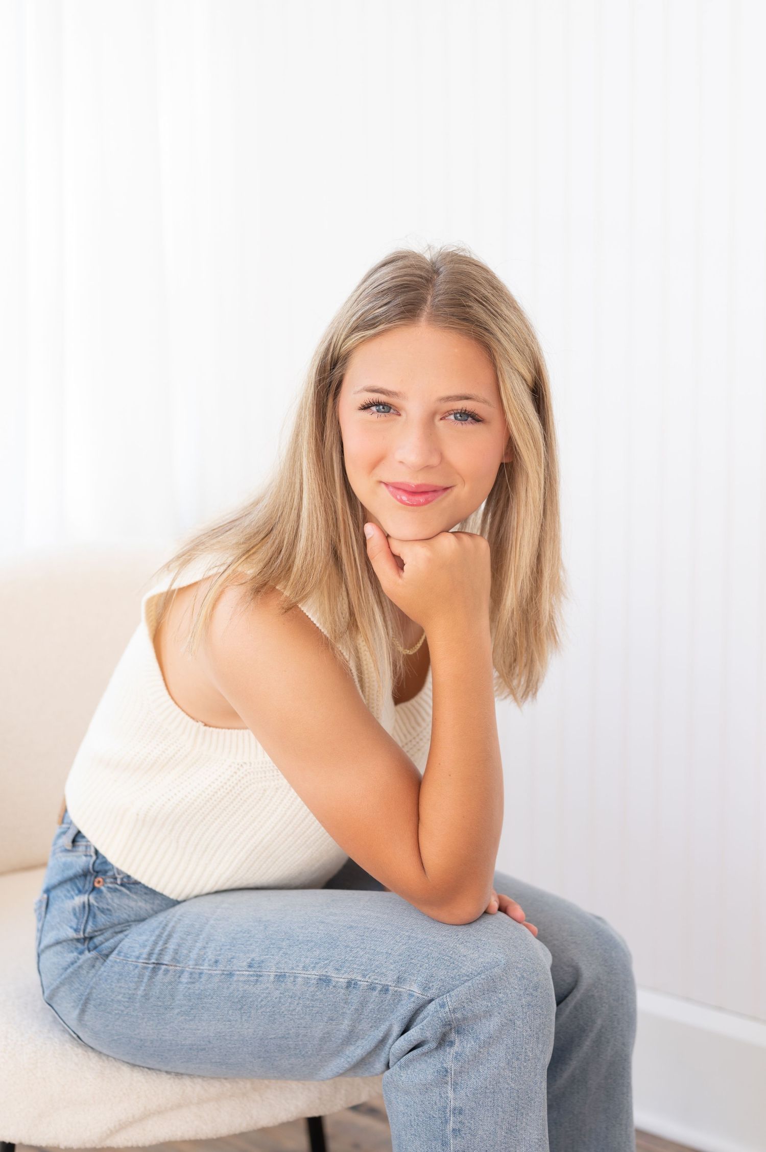 A person wearing a white sleeveless top and blue jeans poses casually against a light background.