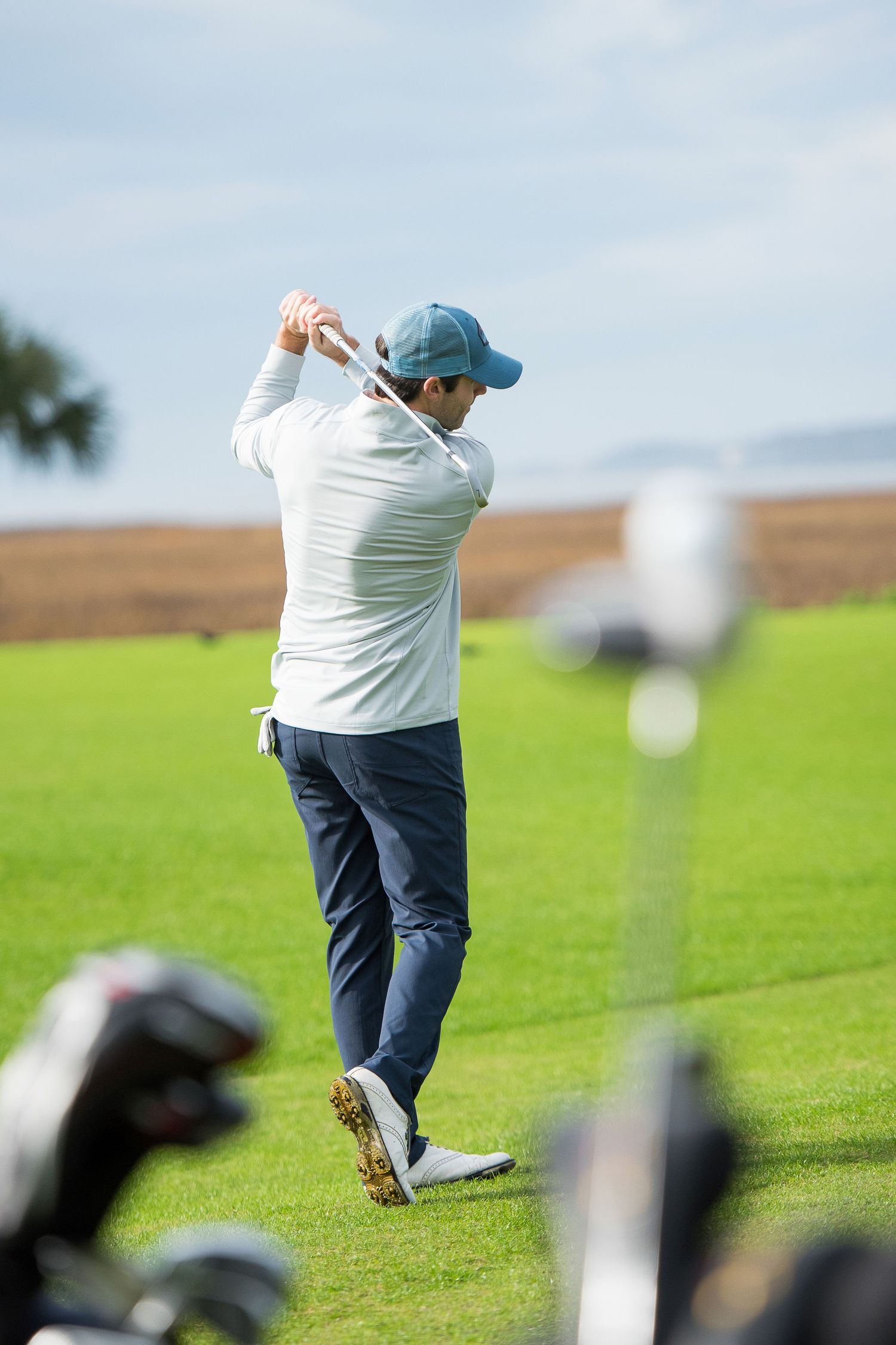 Golfer in a blue cap and white shirt completes a backswing on a sunny green fairy with blurred clubs in the foreground.