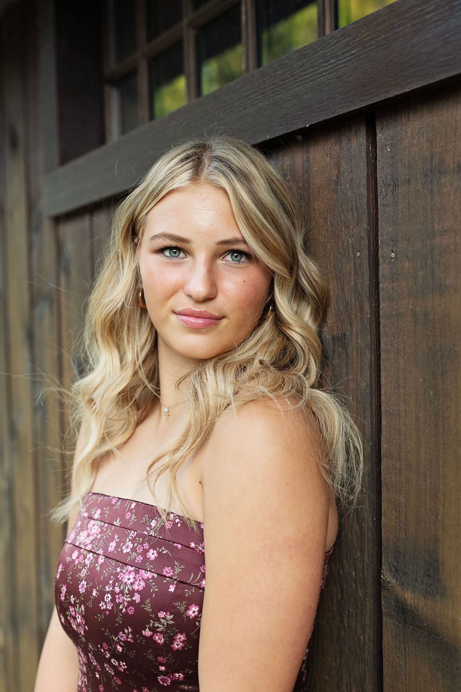 A woman in a floral burgundy dress poses outdoors against a wooden fence background.