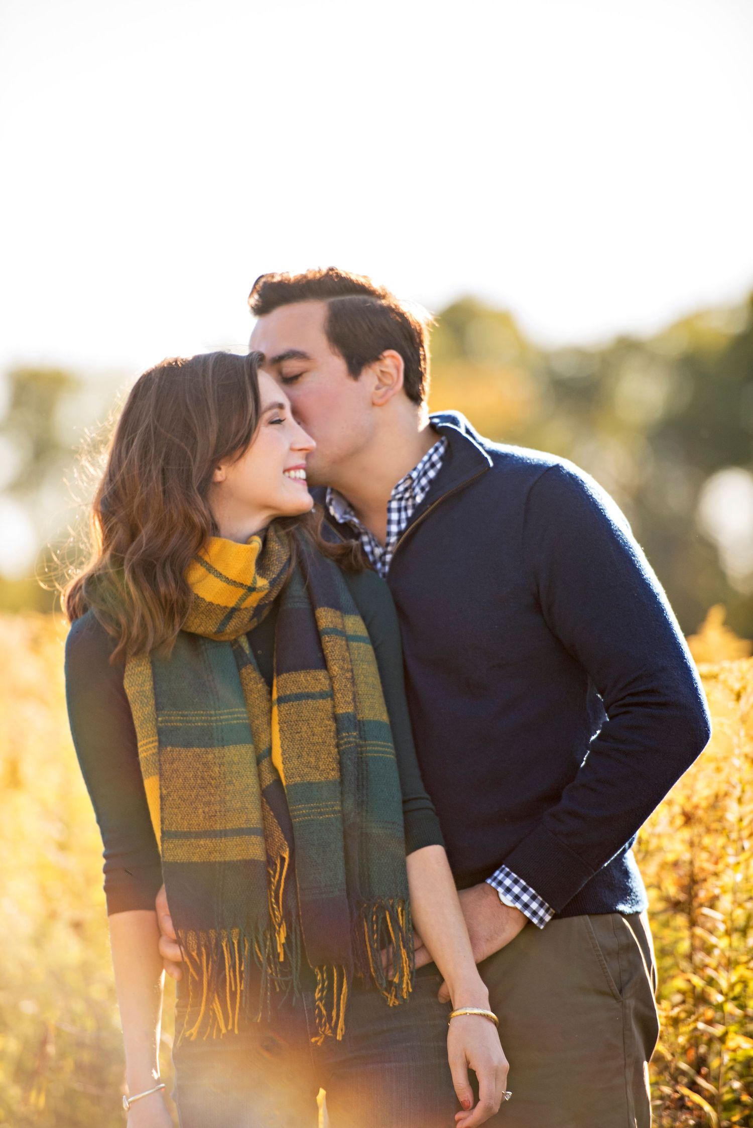 A couple in autumn attire share romantic kisses against a golden sunlit backdrop of fall foliage.