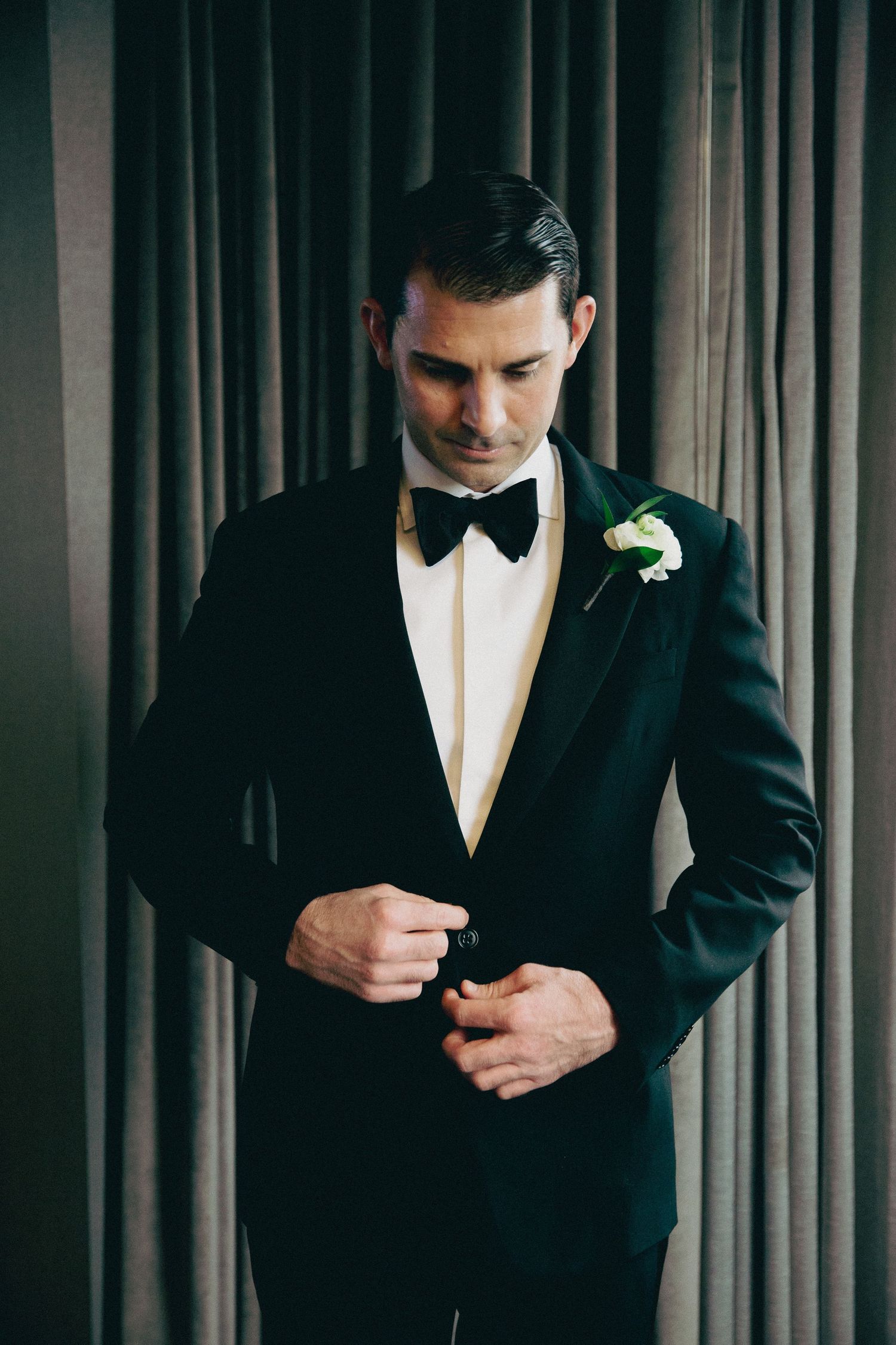 Close-up of groom adjusting black bow tie and tuxedo jacket.