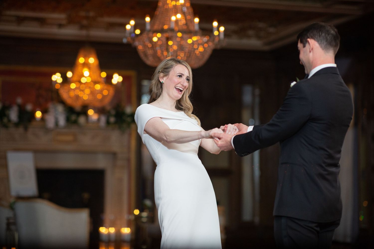 Couple dancing together in an elegant ballroom with ornate chandeliers overhead during a wedding celebration.