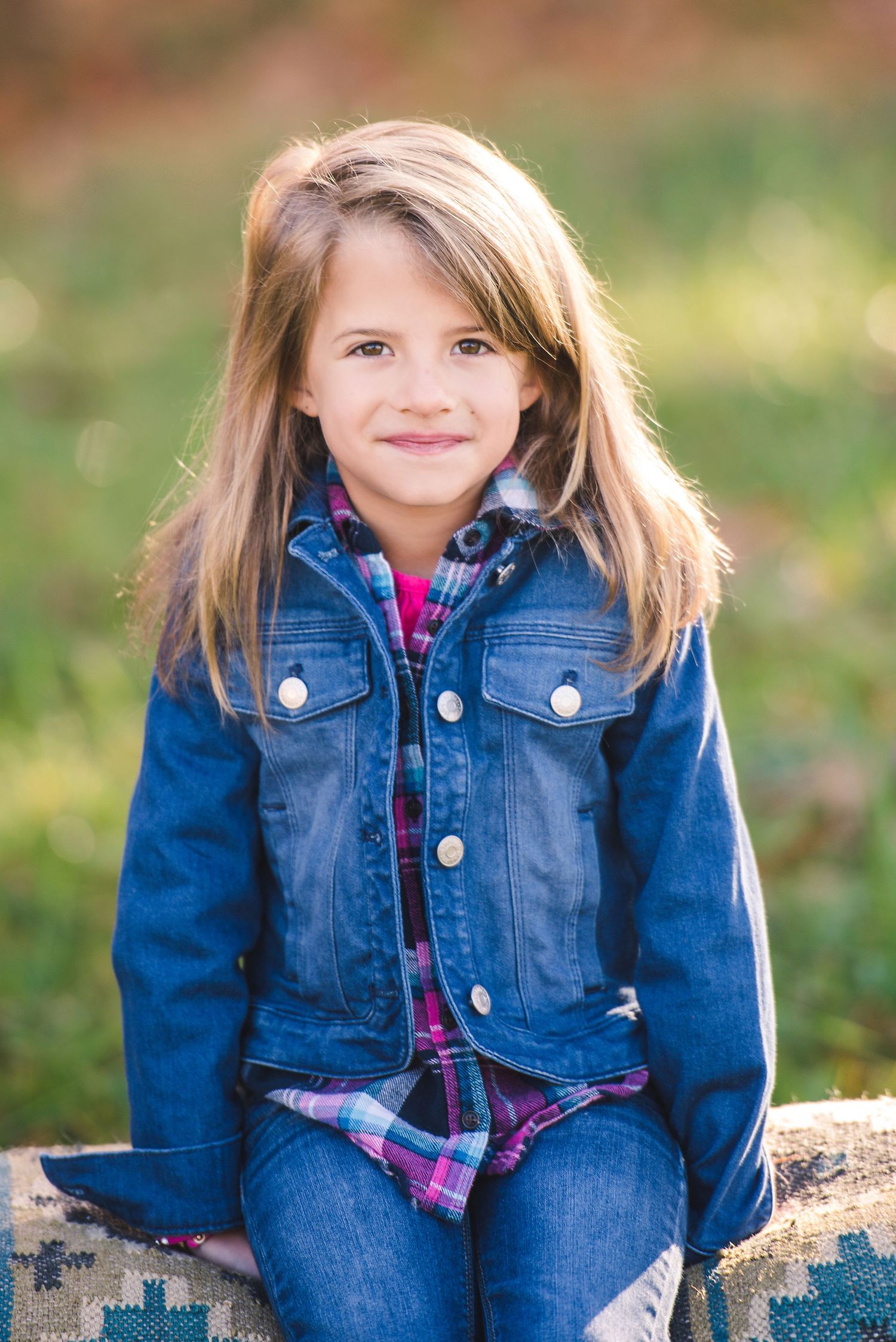 Child wearing denim jacket and plaid shirt poses outdoors on a sunny autumn day.