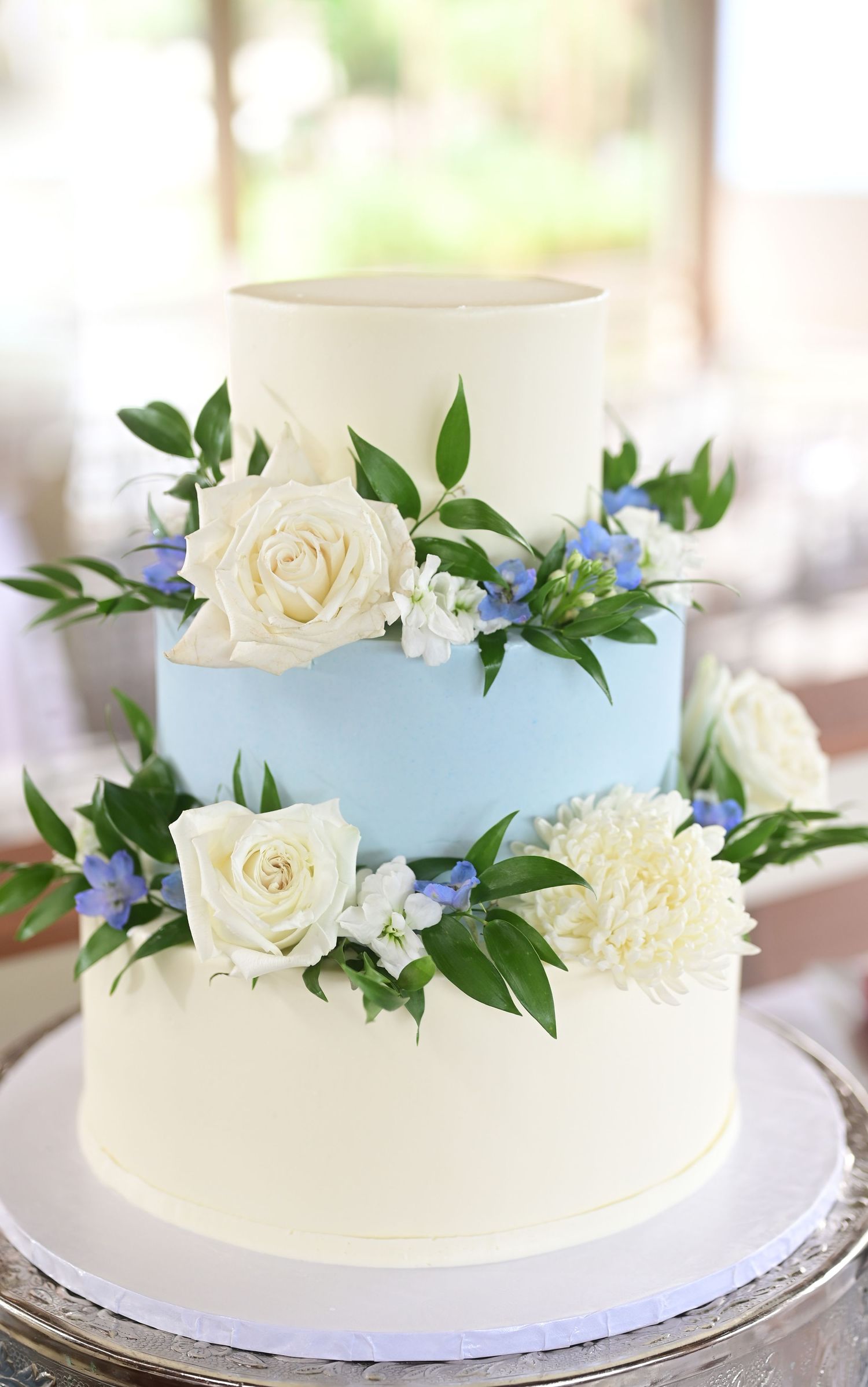 A three-tier wedding cake decorated with white roses and blue flowers on a silver cake stand.