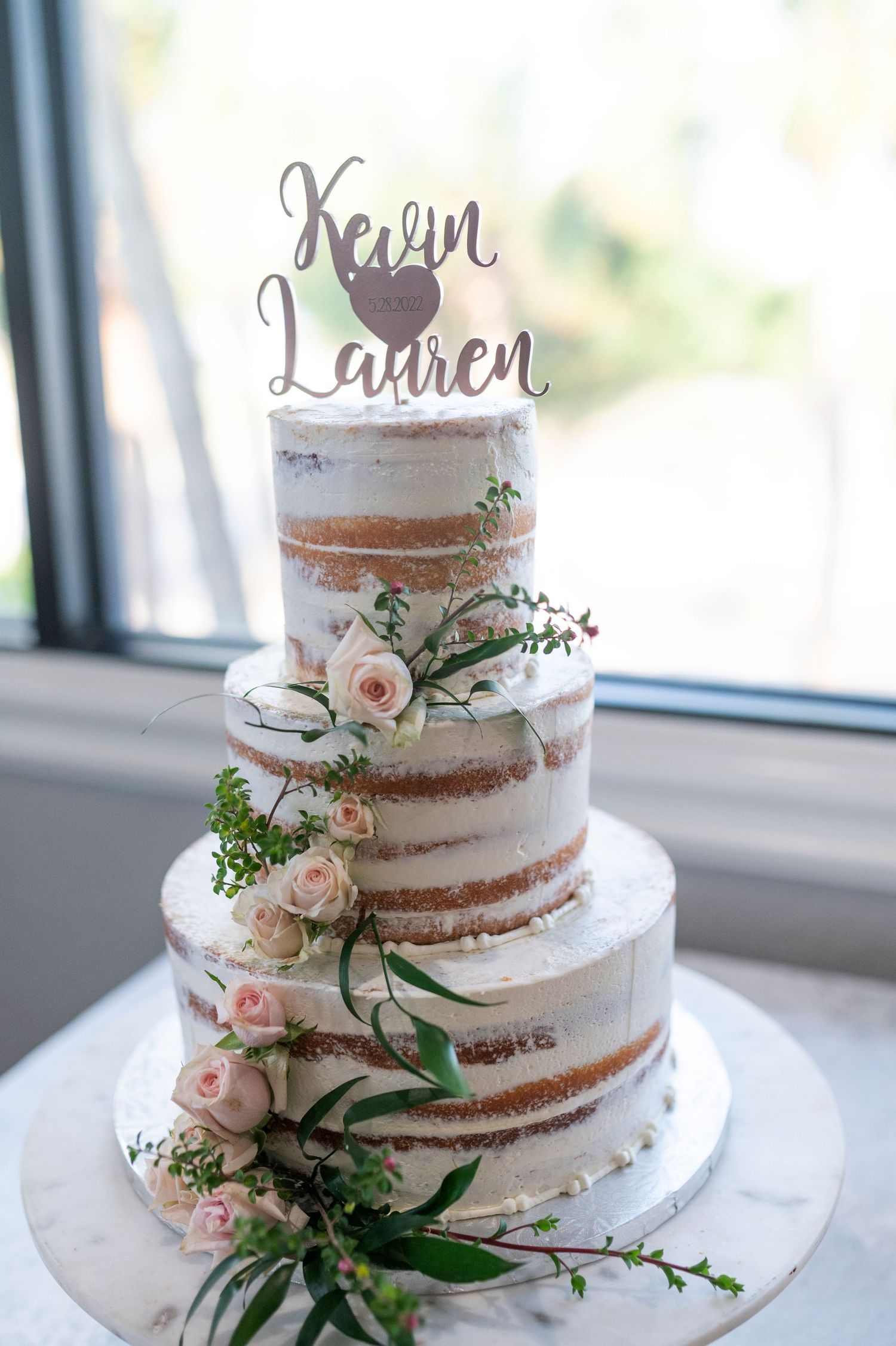 Rustic semi-naked wedding cake decorated with fresh pink roses and greenery on display near a window.