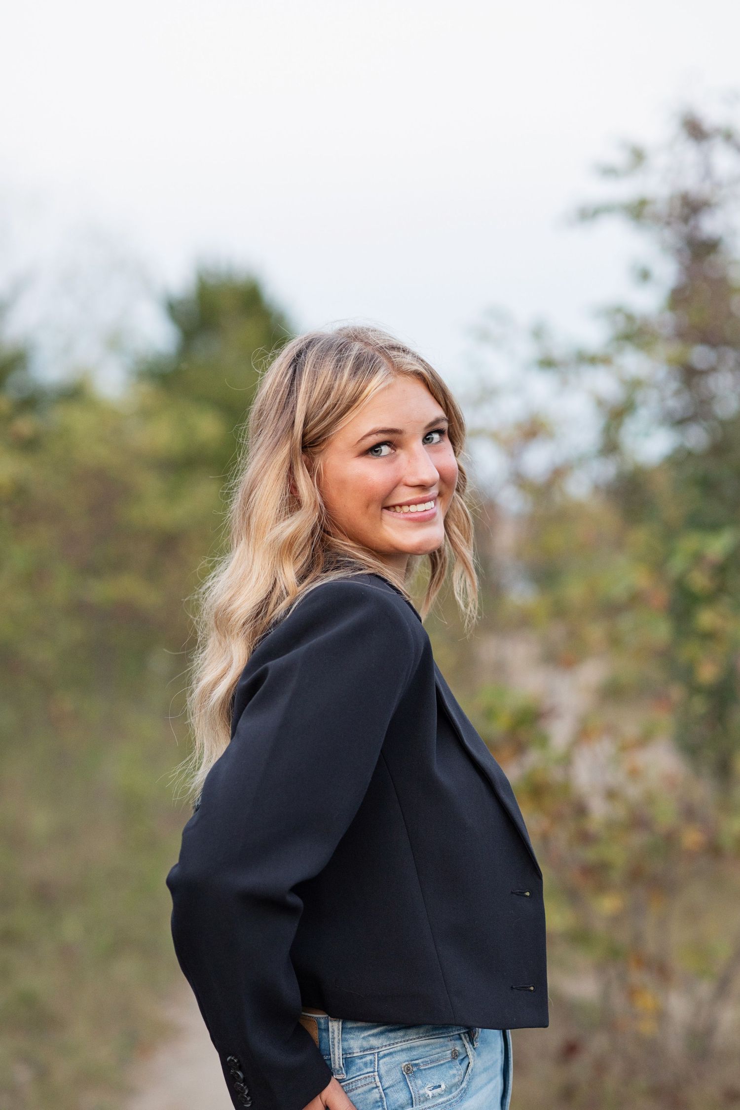 Person wearing a black cropped jacket and blue jeans poses outdoors against a blurred natural background.
