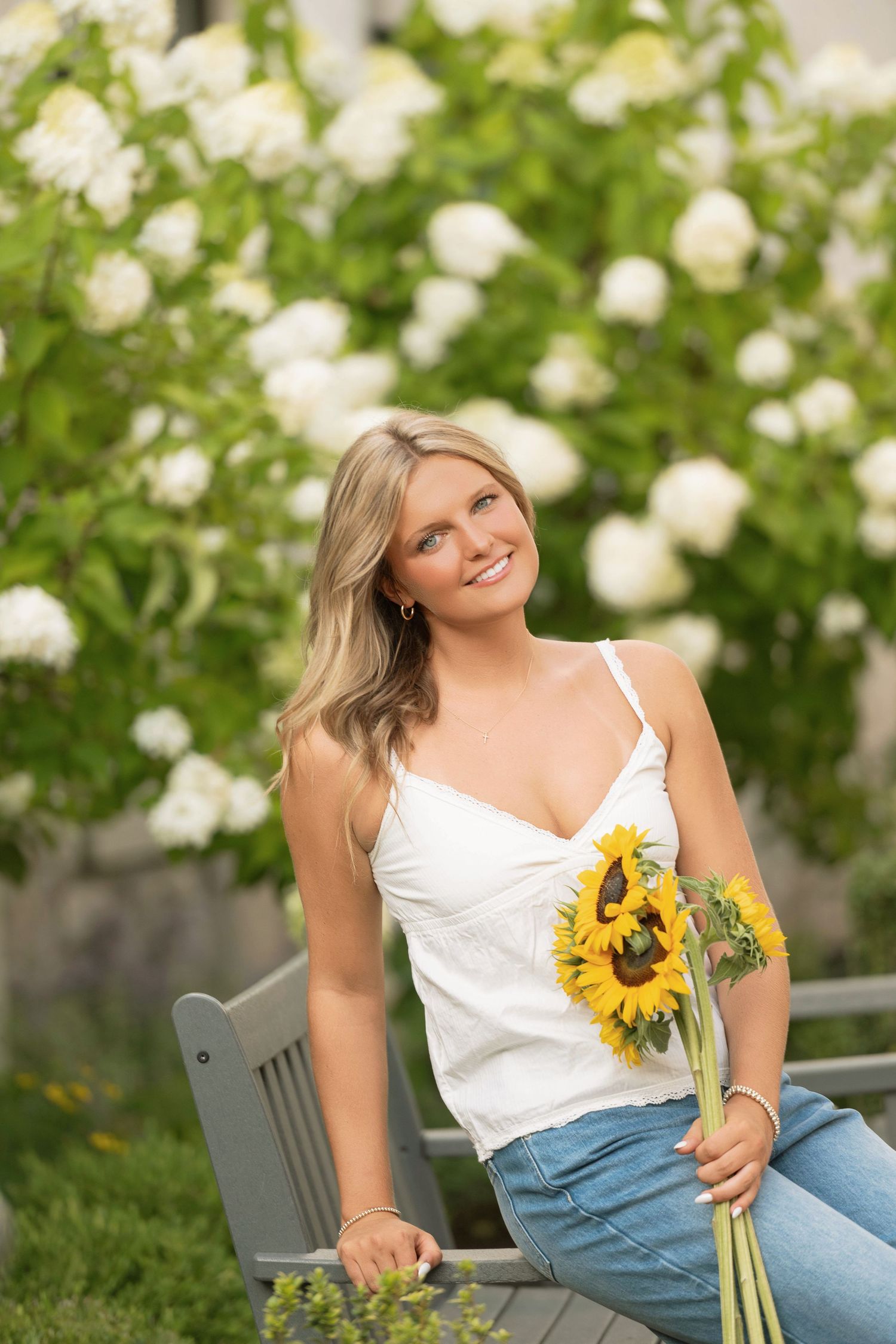 Woman in white top and jeans lounges on bench with sunflowers in garden setting with white roses blooming.