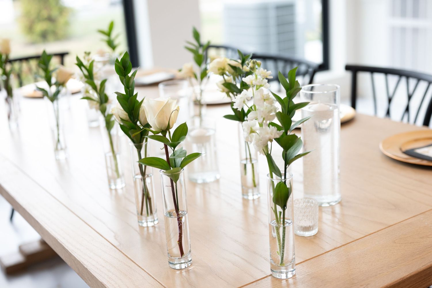 White flowers in slim glass vases arranged on a wooden dining table in a bright modern setting.