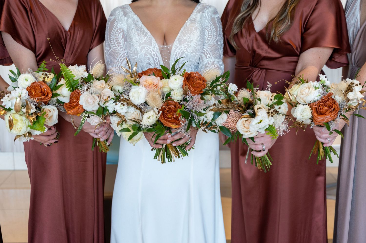 Wedding party holds beautiful bouquets with orange roses and white flowers against white gown and burgundy dresses.