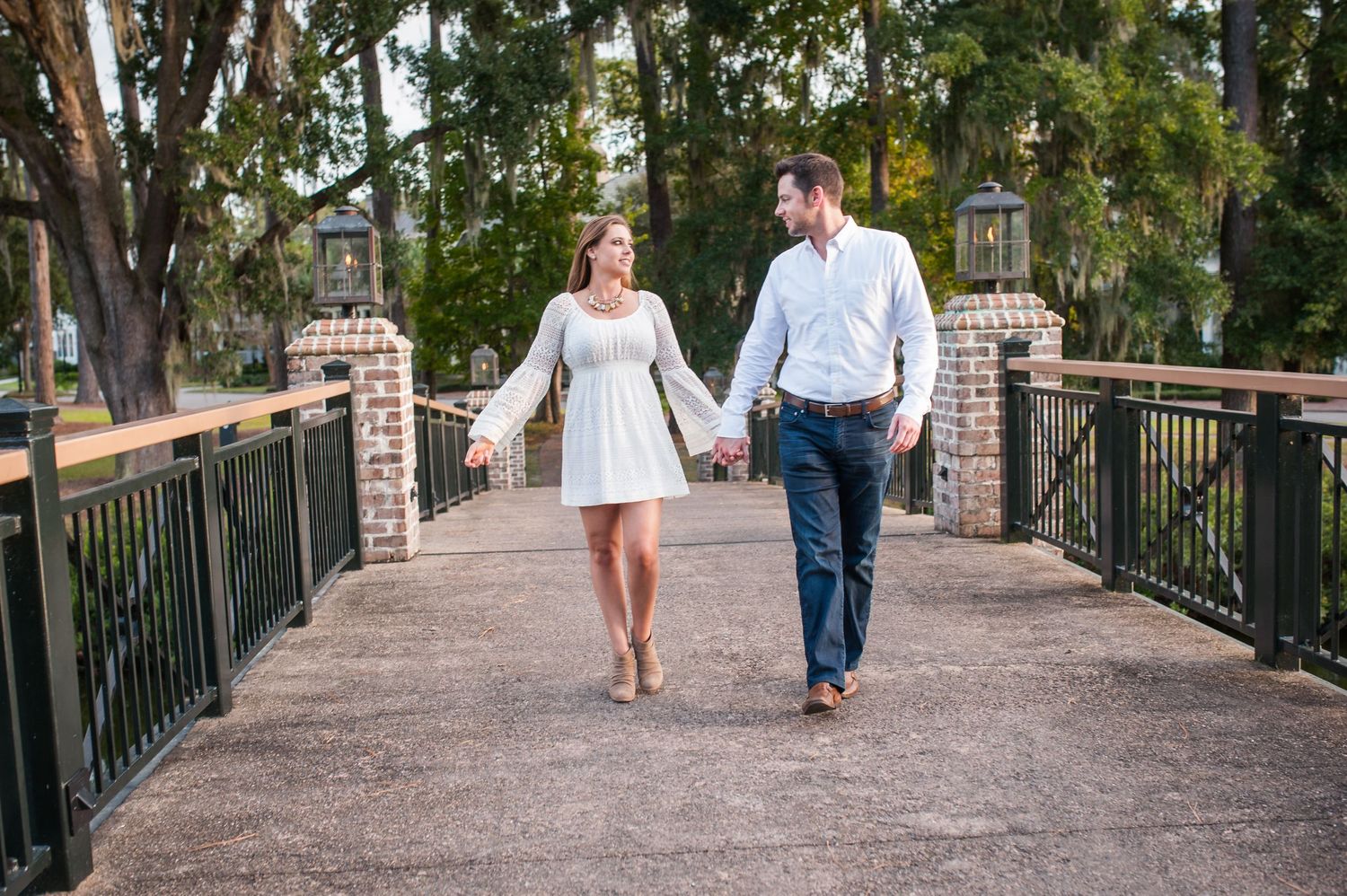 Couple walking hand in hand along a brick pathway beneath towering oak trees.