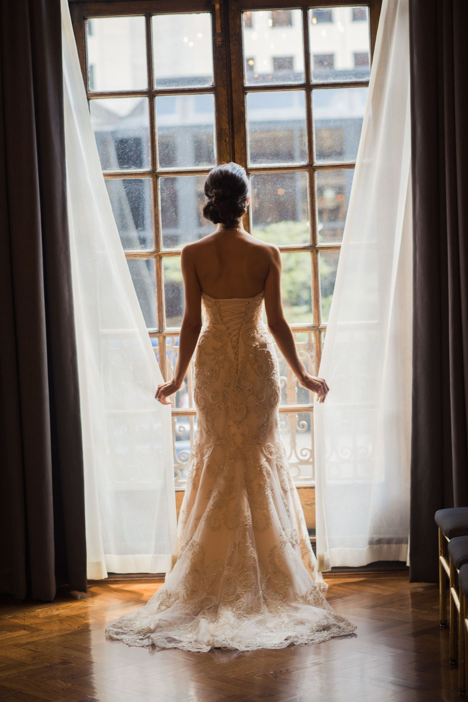 Silhouette of wedding dress against large windows with sheer curtains.