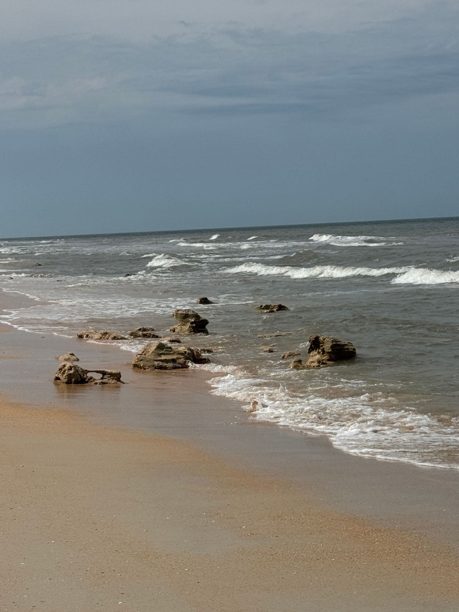 Sandy beach shoreline with rocky outcroppings, gentle ocean waves, and overcast sky on a calm coastal day.