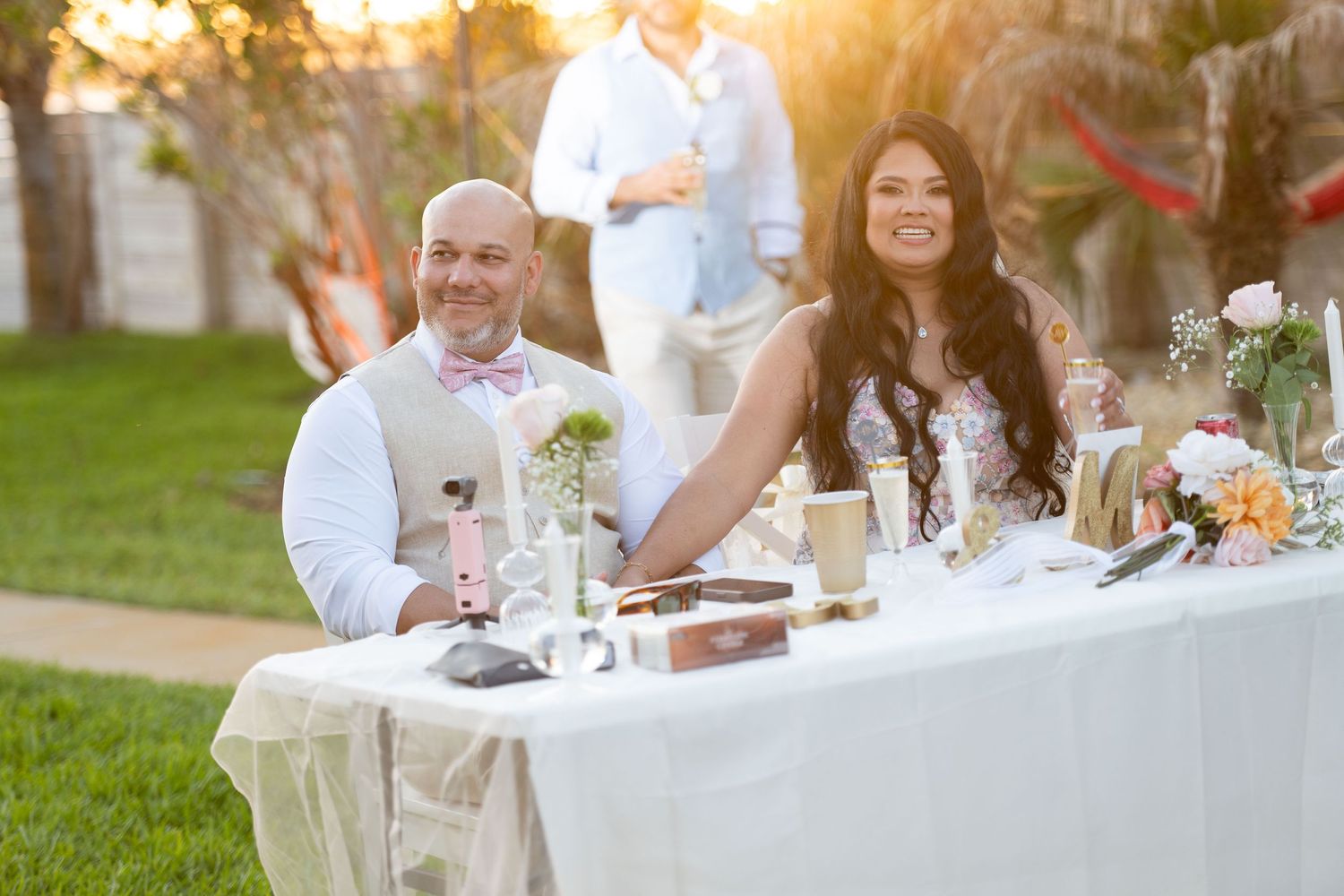 Bride and groom smiling at outdoor wedding reception table with floral centerpieces at golden hour.