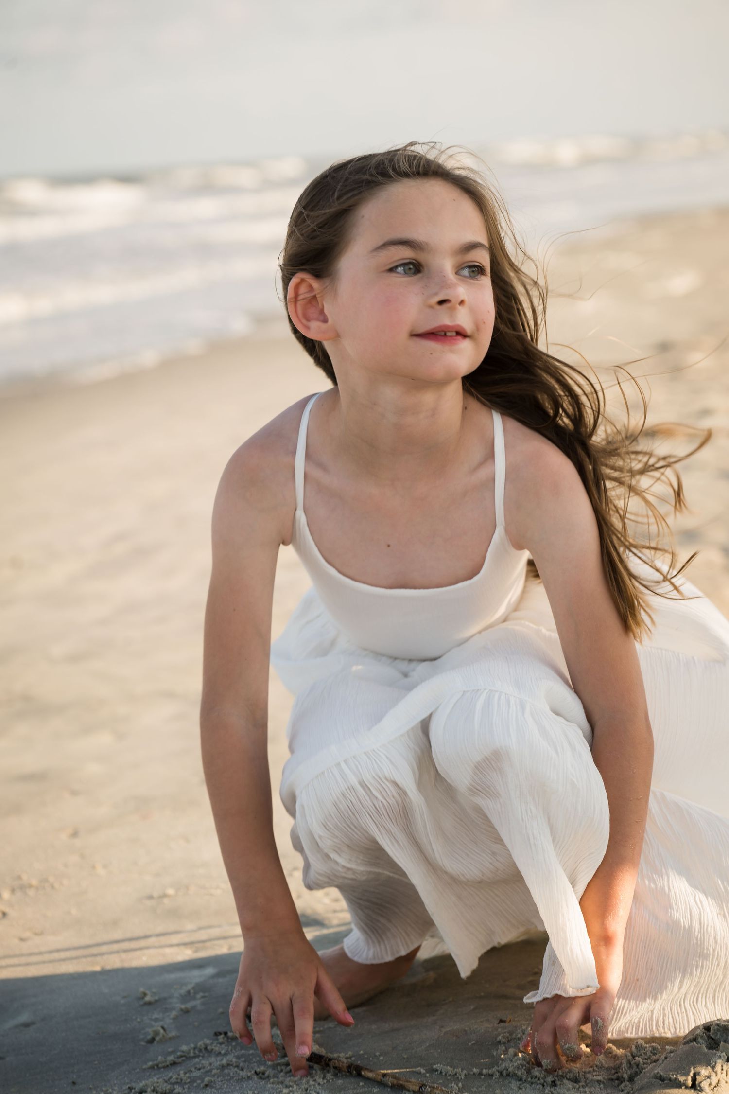 Young girl in white dress crouching on sandy beach with ocean waves in background on a windy day.