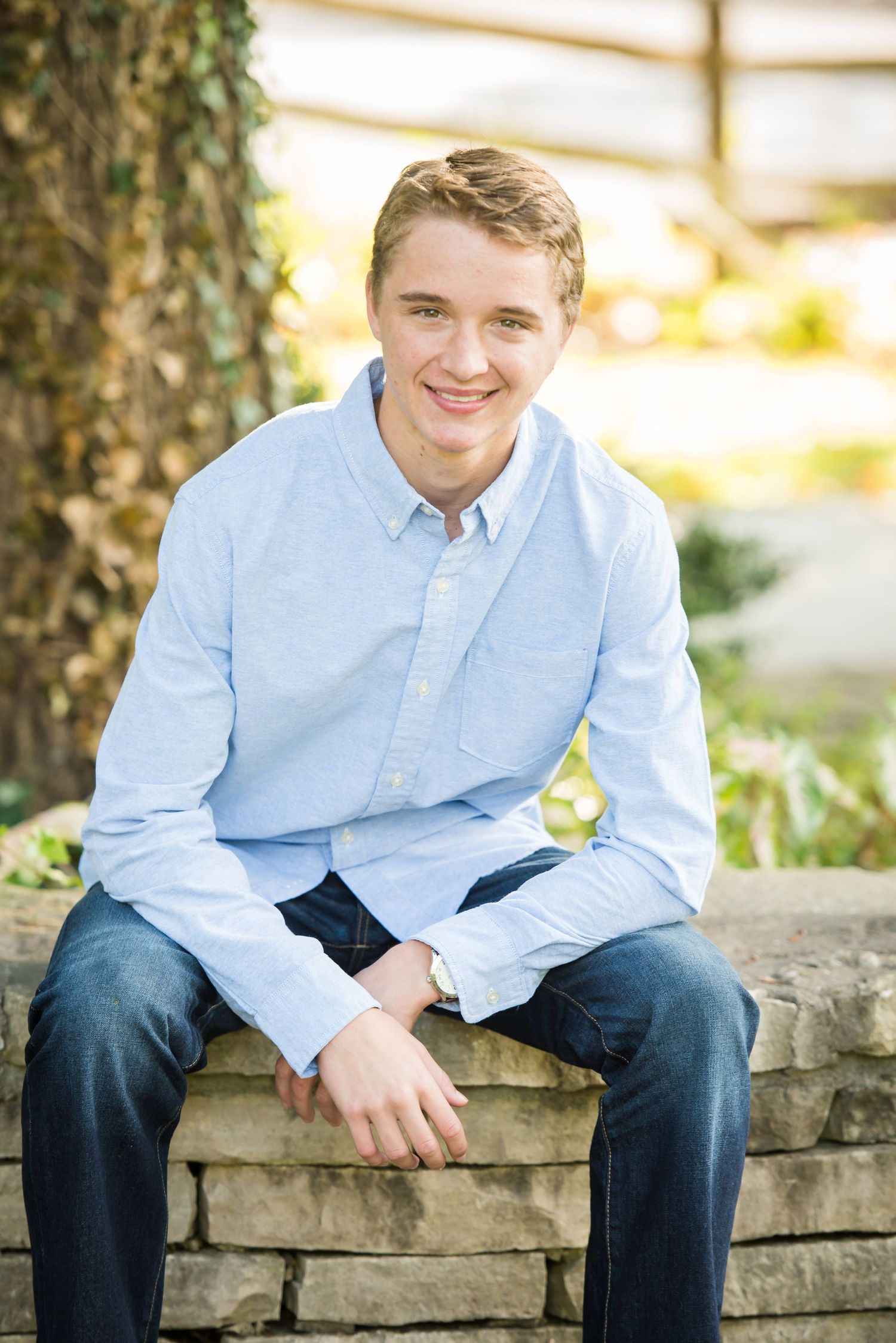 A casual portrait in light blue button-down shirt sitting on stone wall at sunset.