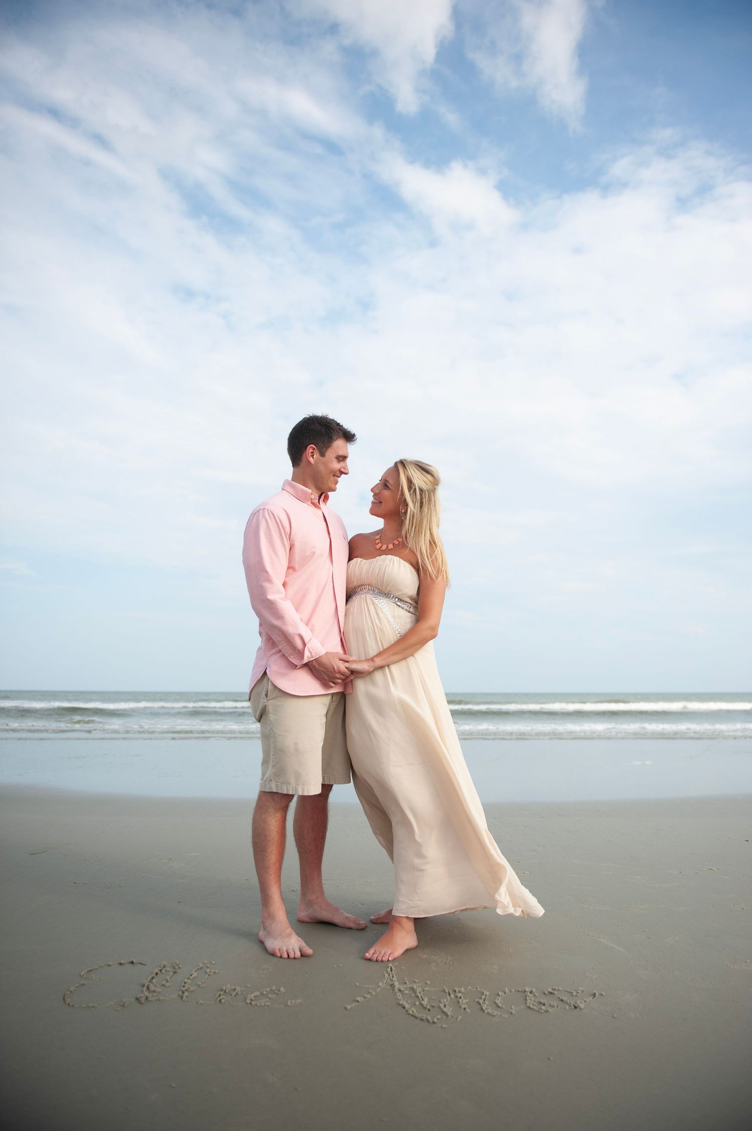 A couple in light colored clothing shares romantic moments on a sandy beach while writing messages in the sand.