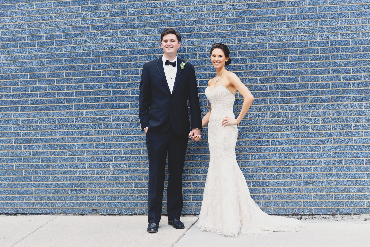 Newlywed couple poses in front of blue brick wall in formal wedding attire.